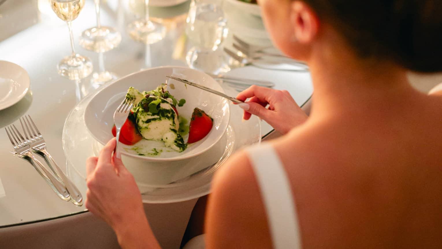 Person in bridal gown sits at round table set for dinner, fork and knife posed to dig into a meal