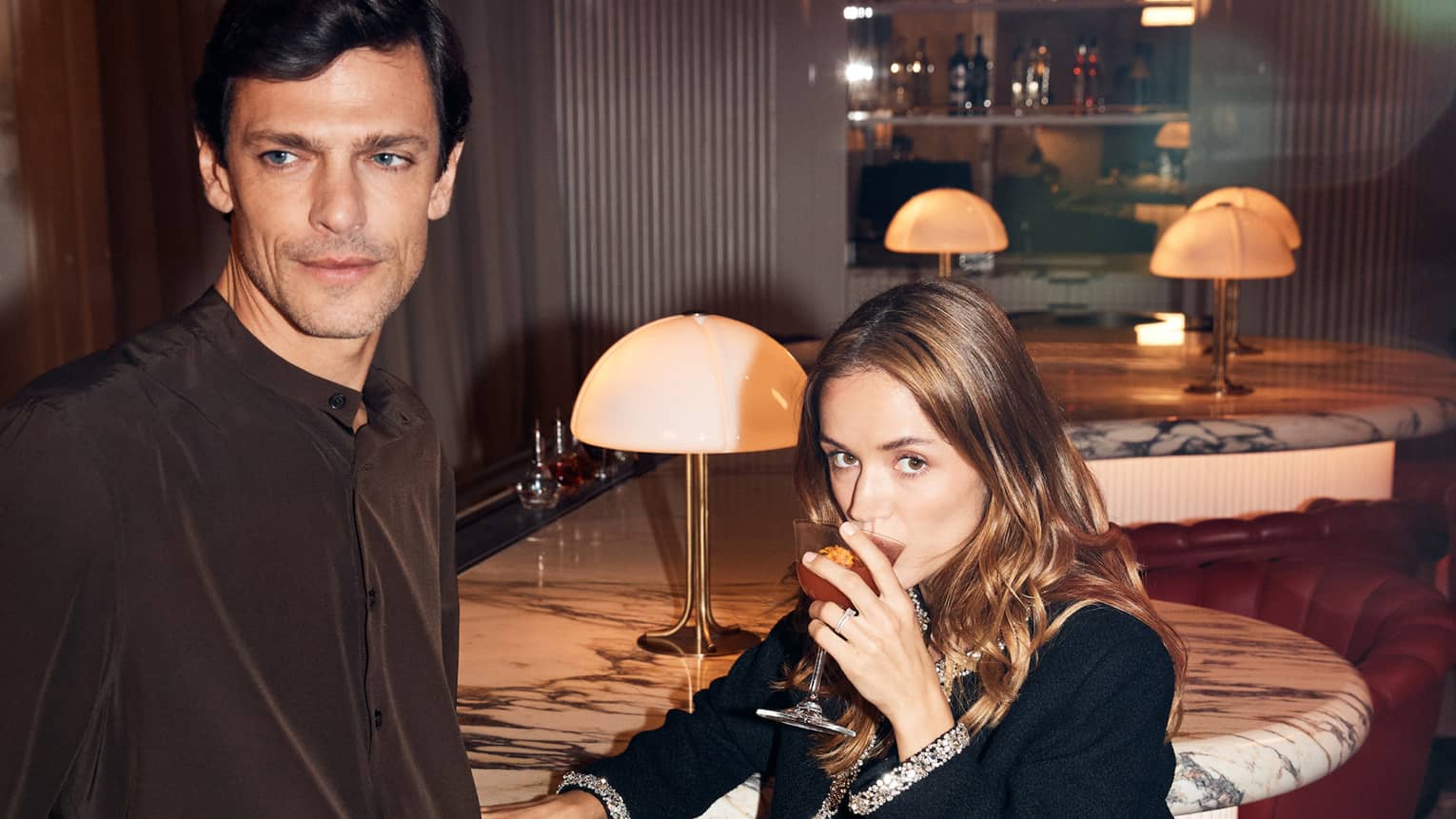 Two people in cocktail attire sit and stand around small red chairs in a dimly lit lounge
