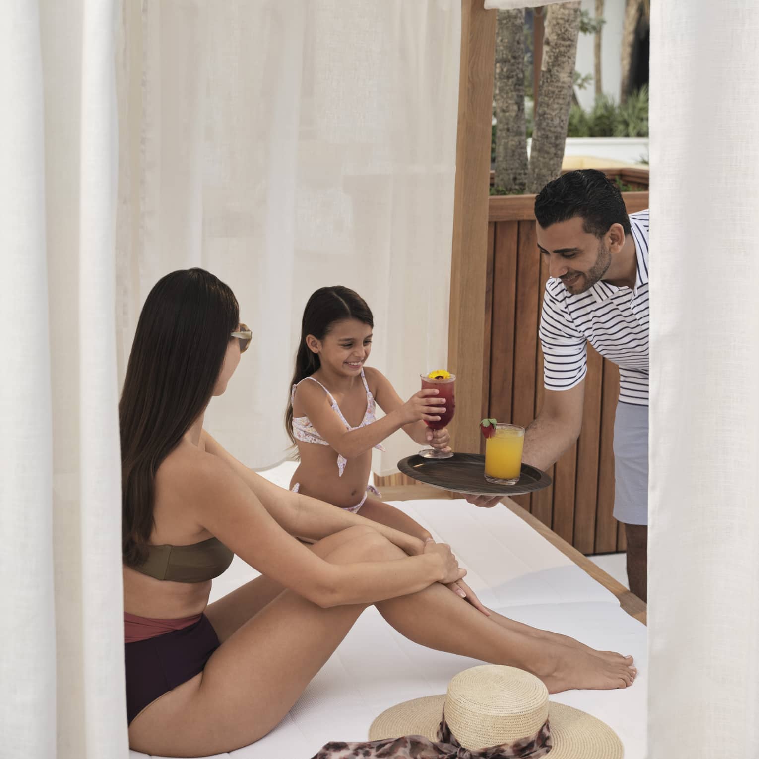 Two guests wearing swimsuits and sitting in a cabana while a person holds a plate with two drinks.