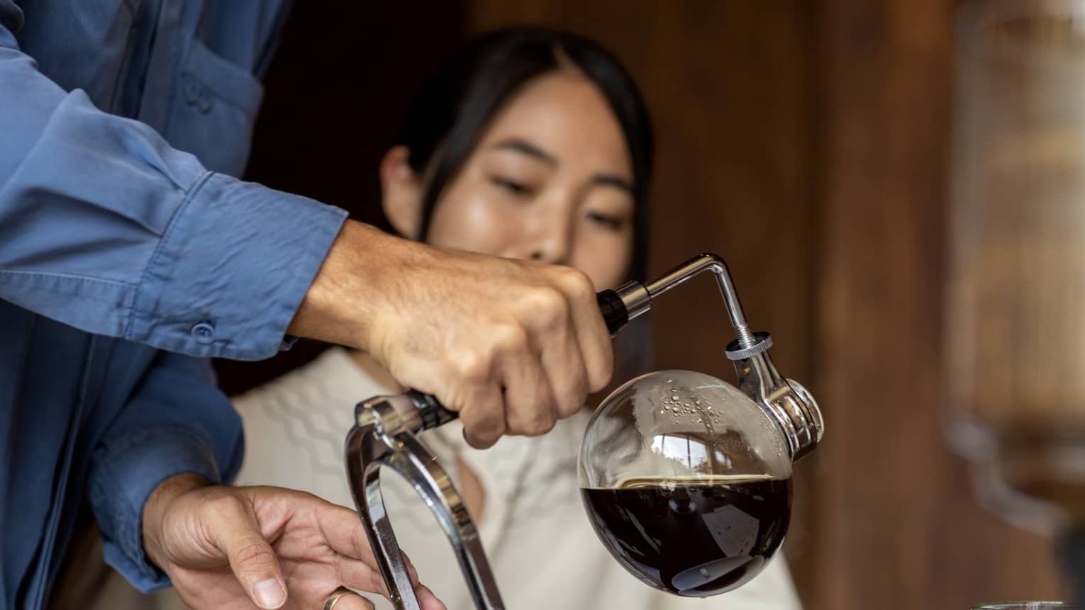 Coffee is poured from a glass bulb attached to a metal stand into white porcelain cups while a guest looks on. 