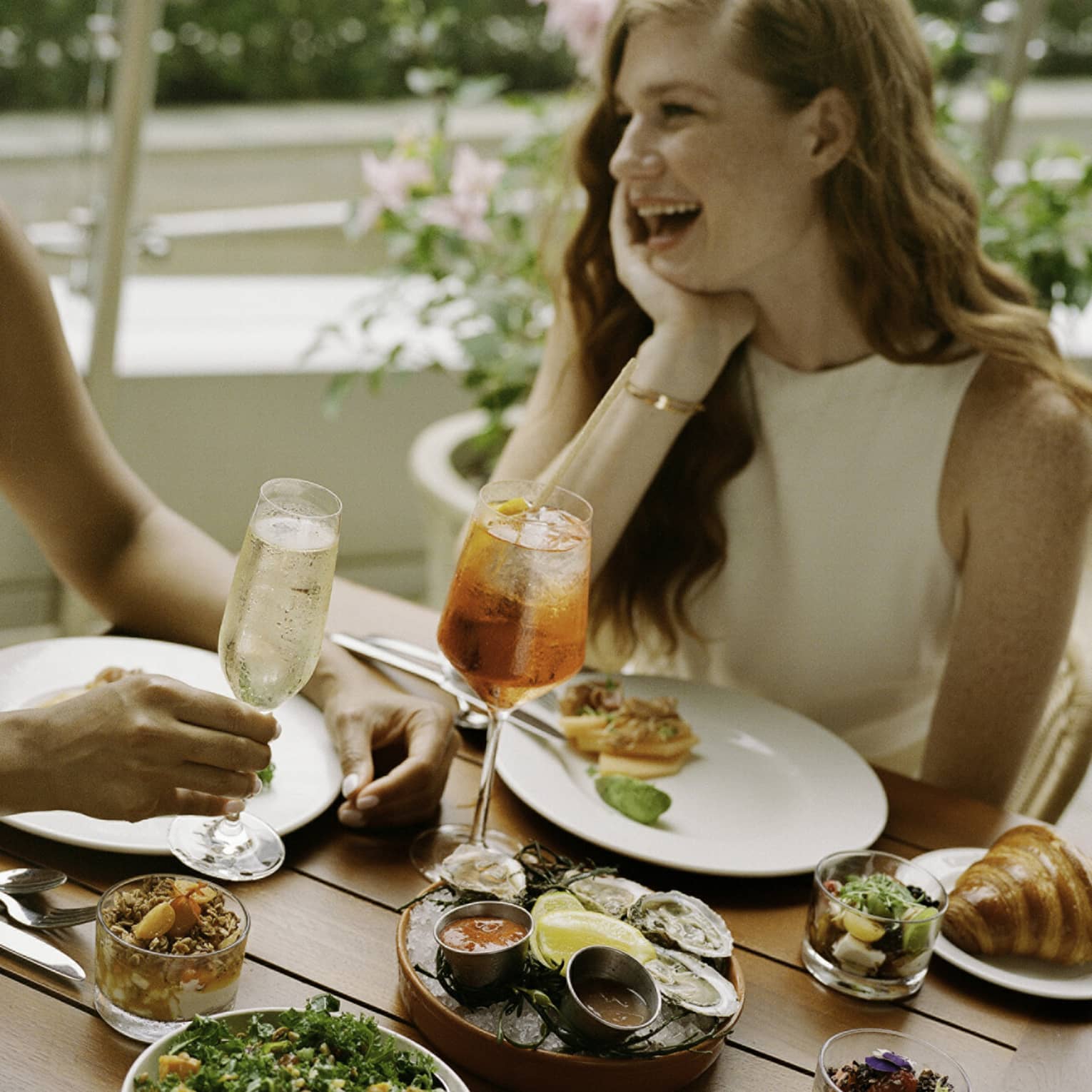 Laughing guests enjoying cold beverages, raw oysters with sauces, smoked salmon, a kale salad, muesli and croissants.