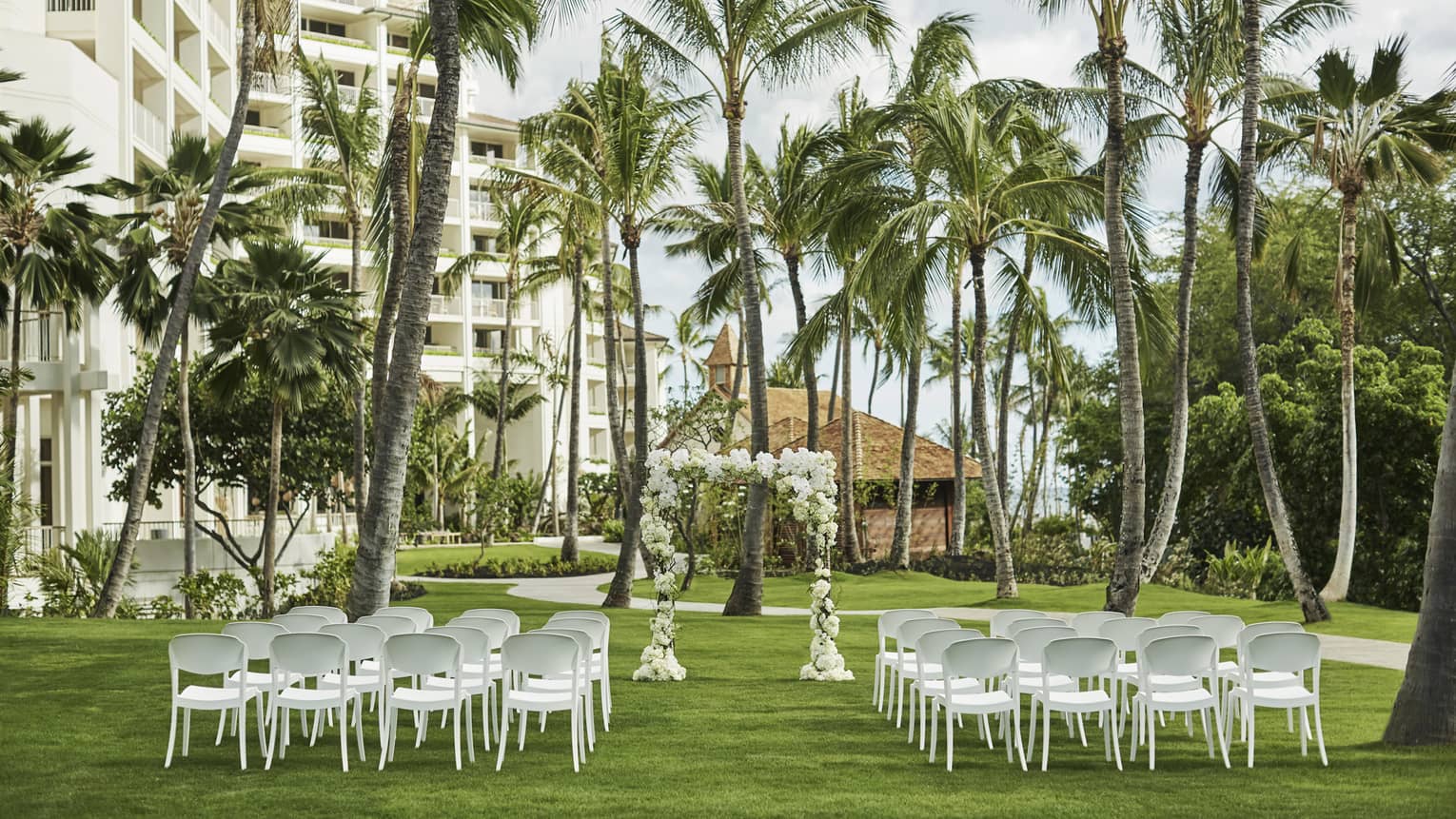 A picturesque outdoor wedding venue with elegant floral arrangements, white chairs on a lush green lawn, beneath palm trees