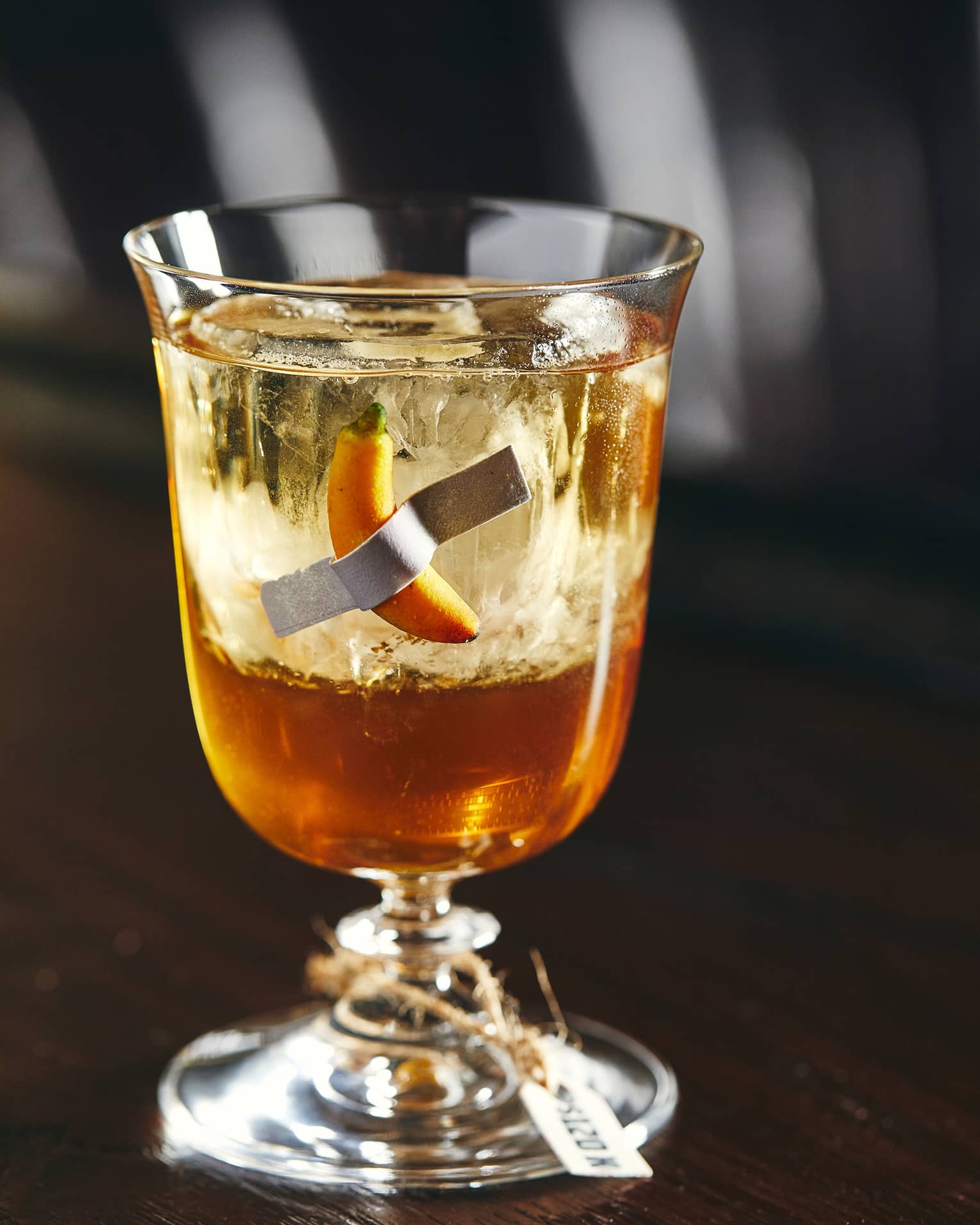 An amber coloured cocktail with a small yellow pepper floating in it.