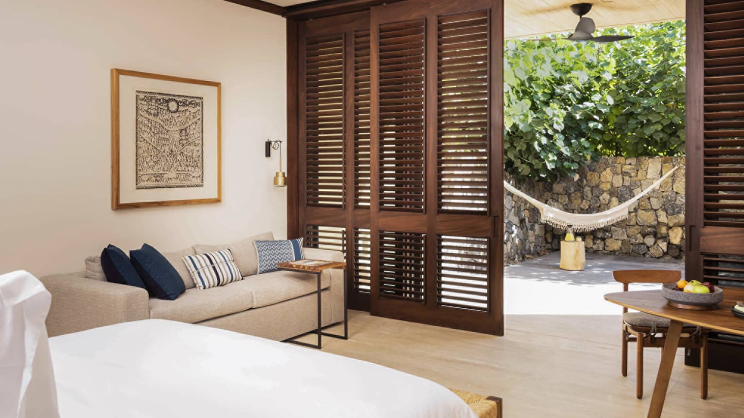 Cozy bedroom with a beige sofa and blue accent pillows, opening to a private patio through wooden shutters. The outdoor space features a hammock and lush greenery.