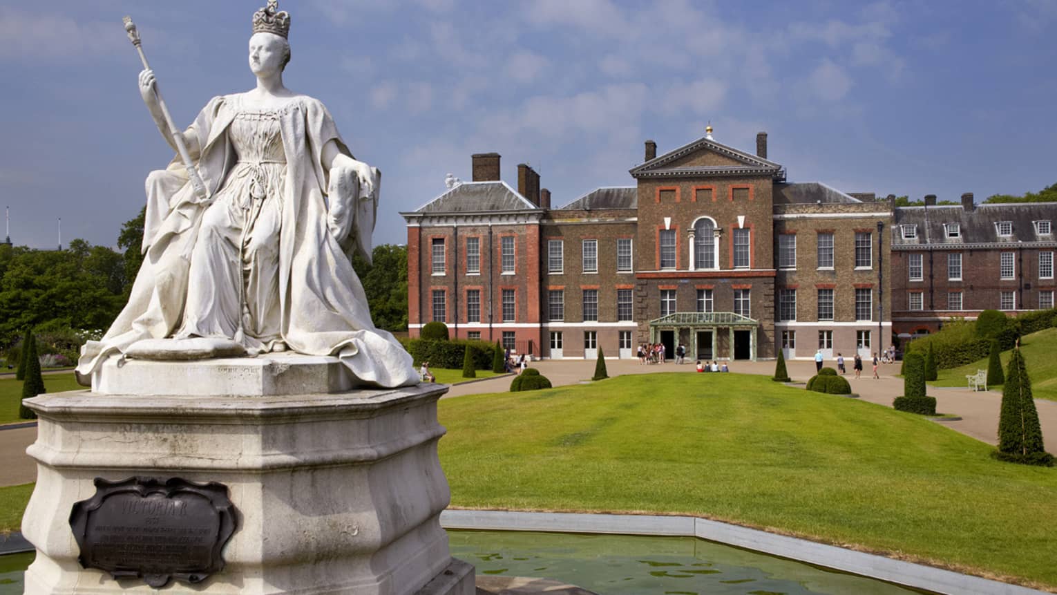 Stately red-bricked Kensington Palace with large green front lawn and white queen statue in forefront