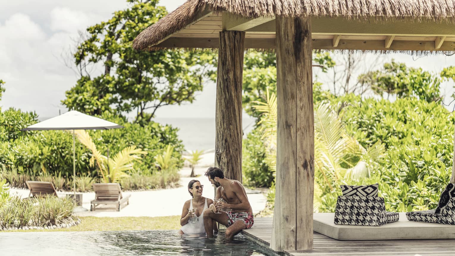 Couple in villa pool under thatched roof and view of tropical foliage