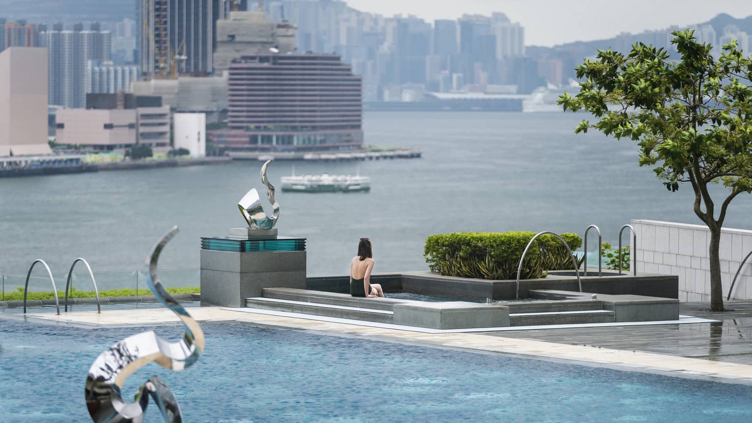A person is seated at the edge of an outdoor swimming pool, which overlooks a large body of water with city buildings in the background. 