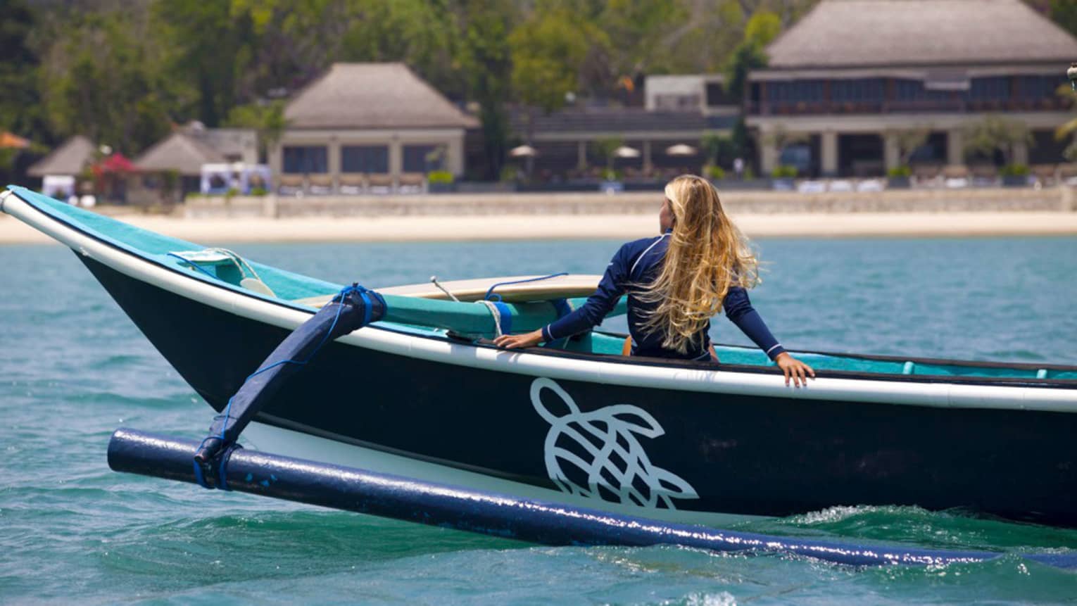 Rear view of a guest reclining in an outrigger canoe, surfboard set down nearby, and gazing toward the lush Resort shoreline.