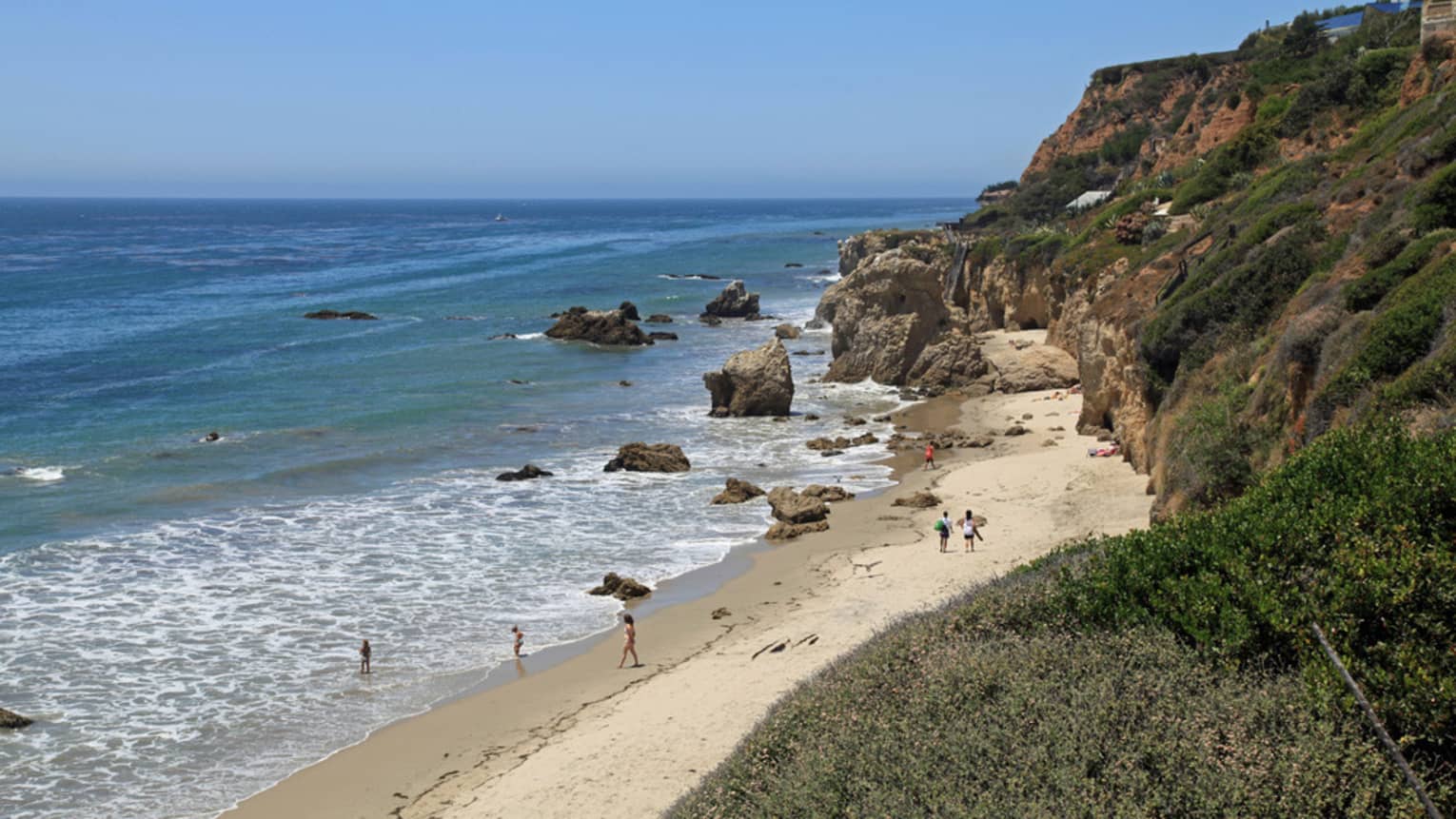 View of people on in ocean, walking on sand, rock formations and shrubbery 
