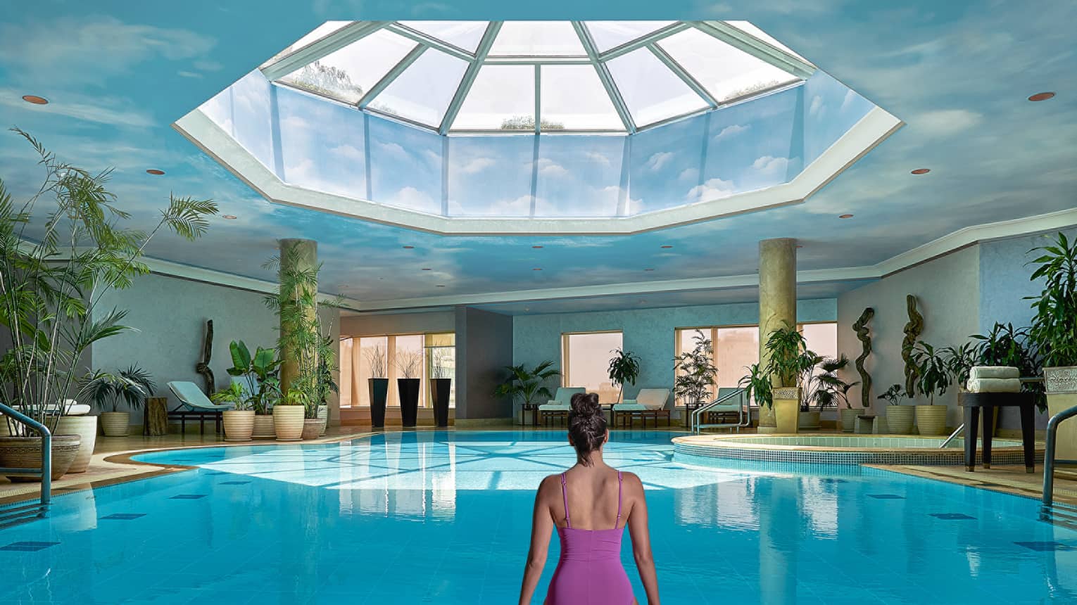 Woman in swimsuit seated on edge of indoor pool with blue ceiling and skylight above