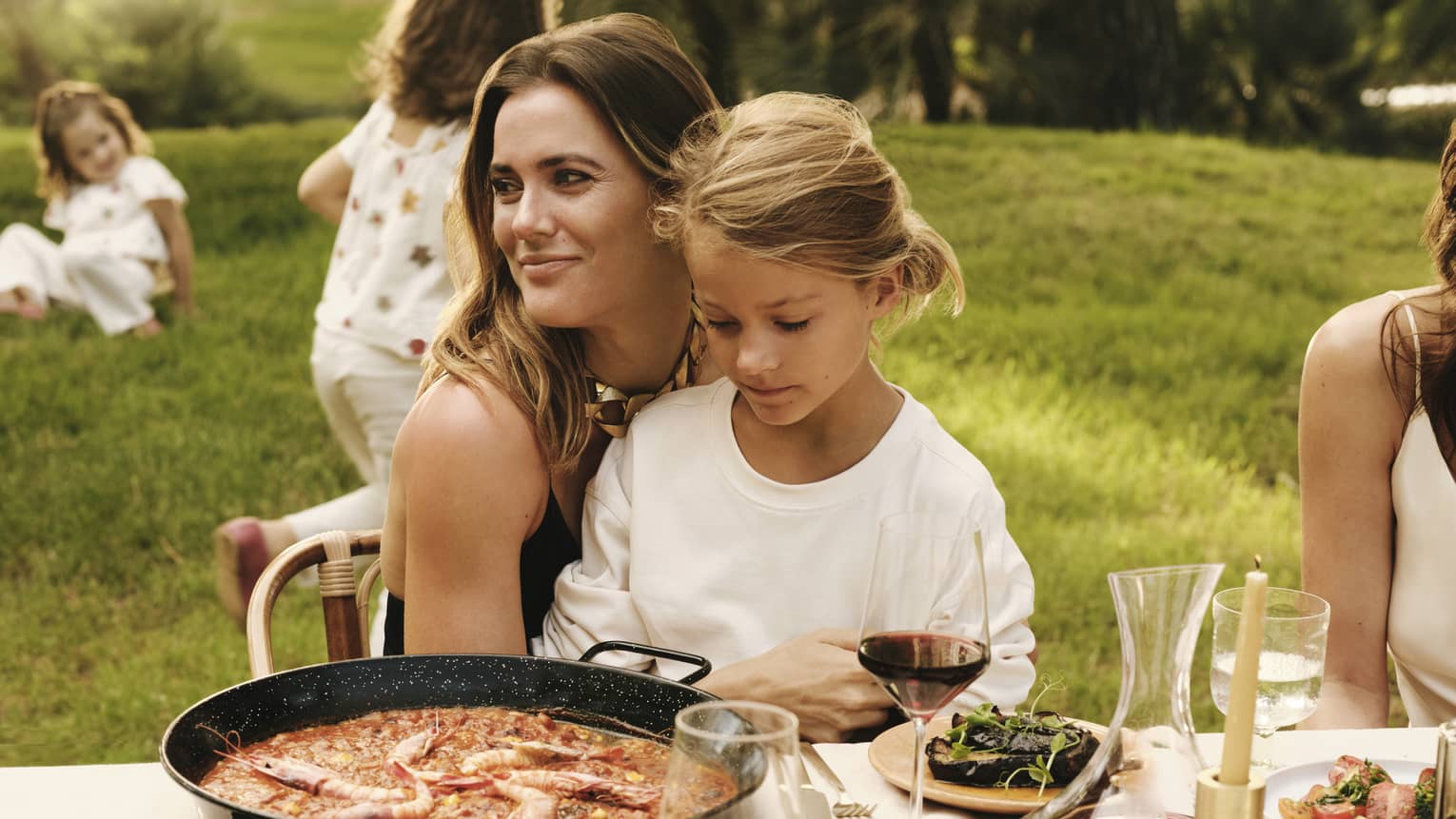 A smiling guest and a child sit together at an outdoor dining table set with a pan of paella, fresh seafood dishes and wine.