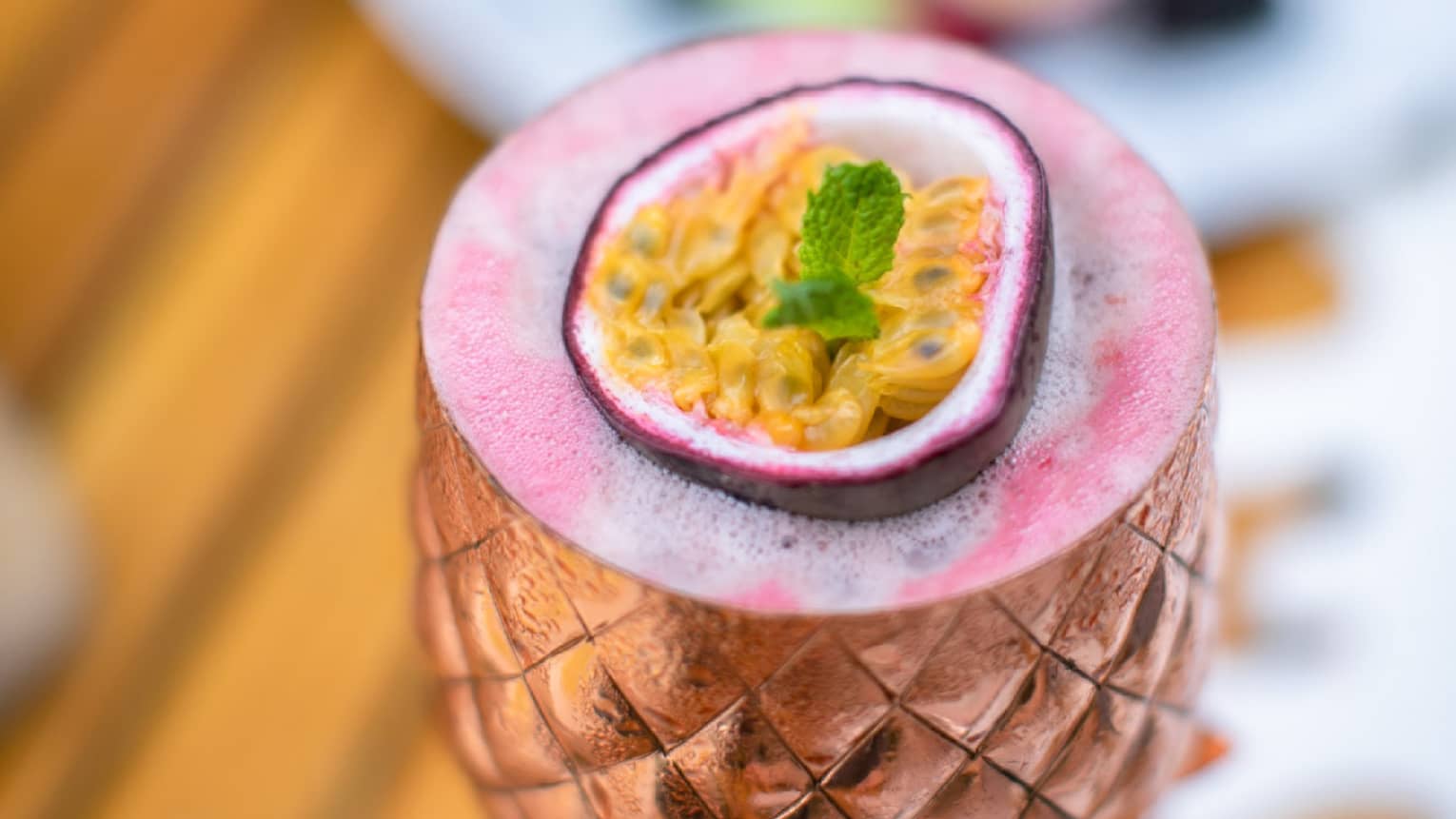 Pink cocktail in bronze pineapple-shaped goblet, garnished with passion fruit