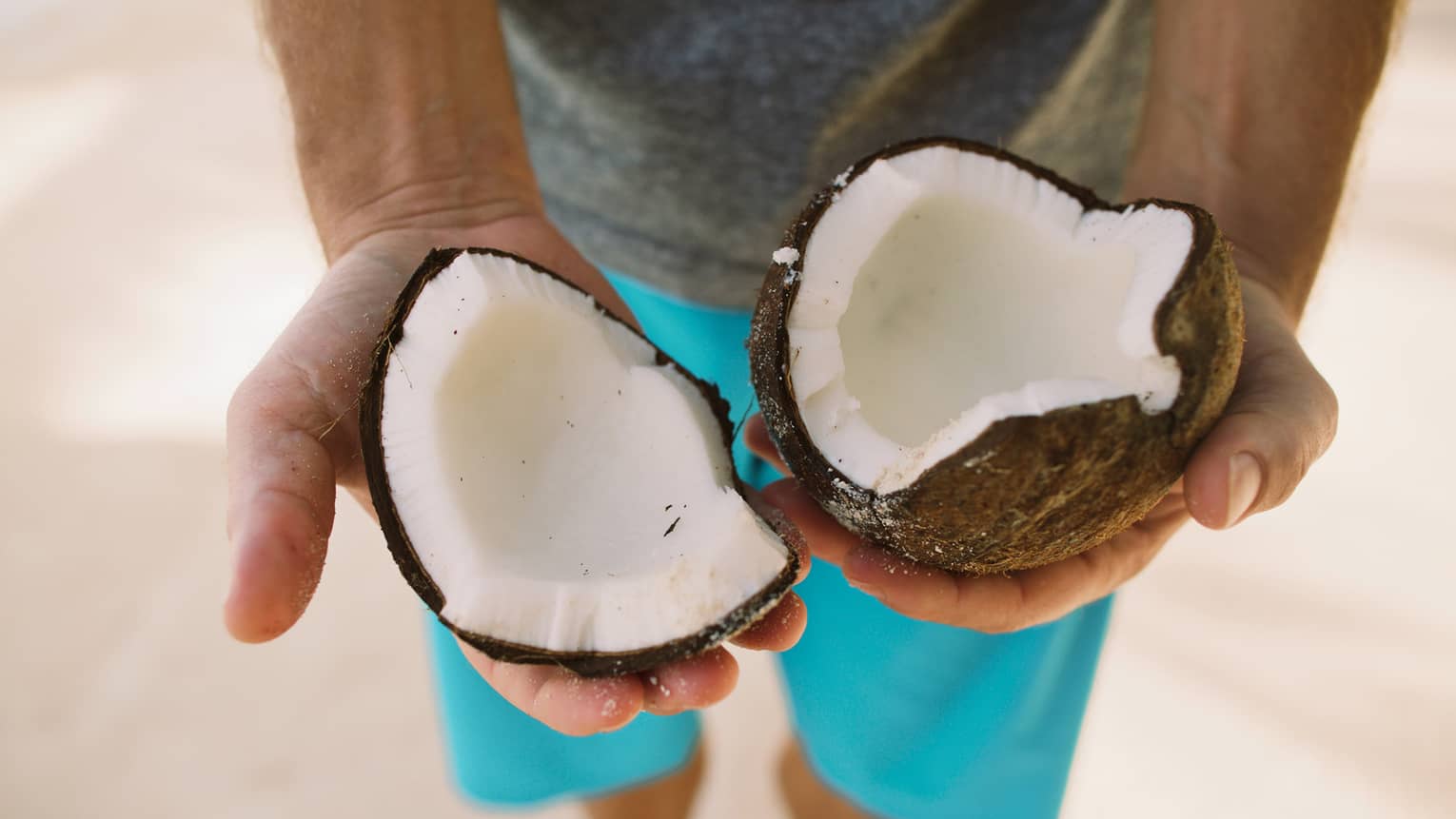 Closeup of two halves of a broken-open coconut showing white meat inside, held by someone in bright turquoise shorts.