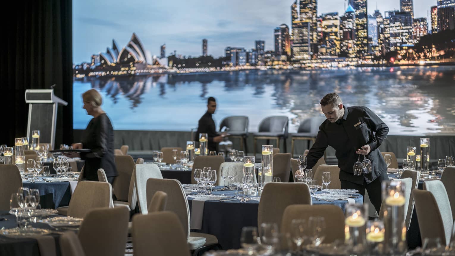 Hotel staff set glassware on banquet tables in ballroom, skyline projection on wall