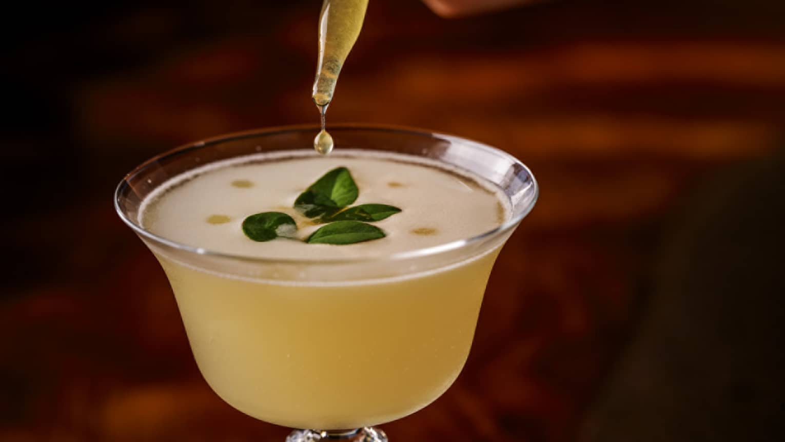 Eye dropper drips yellow liquid into frothy yellow cocktail with green herb garnish