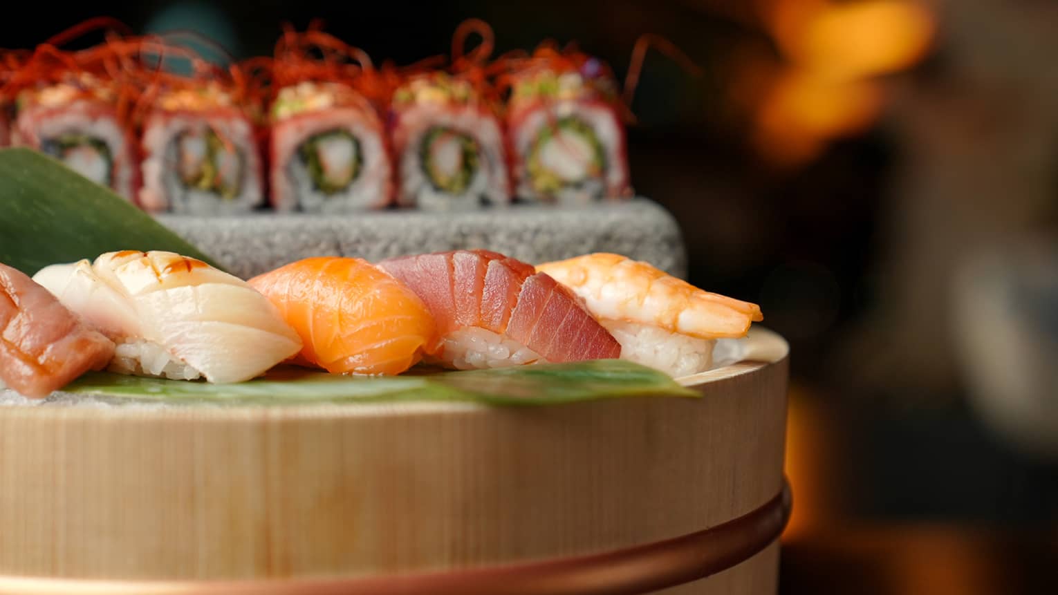 Five pieces of nigiri set on a bamboo platter with more sushi rolls in the background