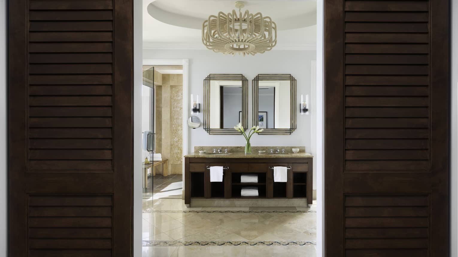 Elegant beachfront villa residence bathroom with double vanity, framed mirrors, and a chandelier, viewed through dark wooden doors