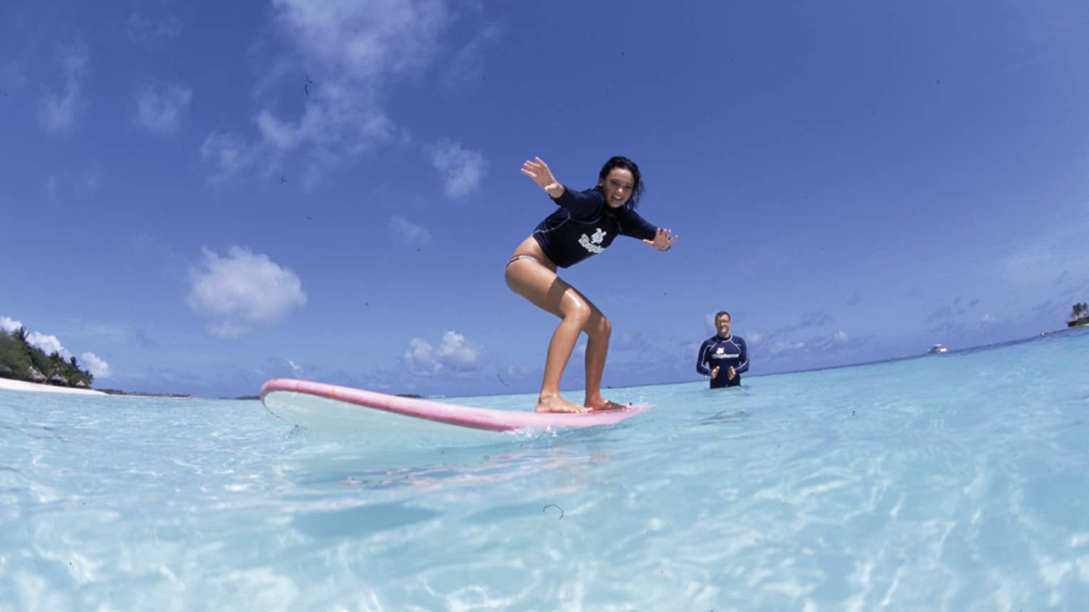 In the crystal-clear shimmering water is a woman learning to surf and an instructor standing in waist-deep water clapping.