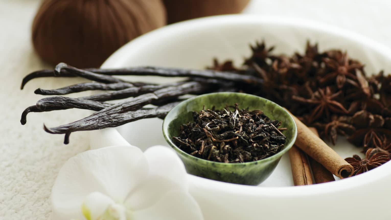 A white bowl filled with various spa ingredients like vanilla bean, star anise and cinnamon sticks