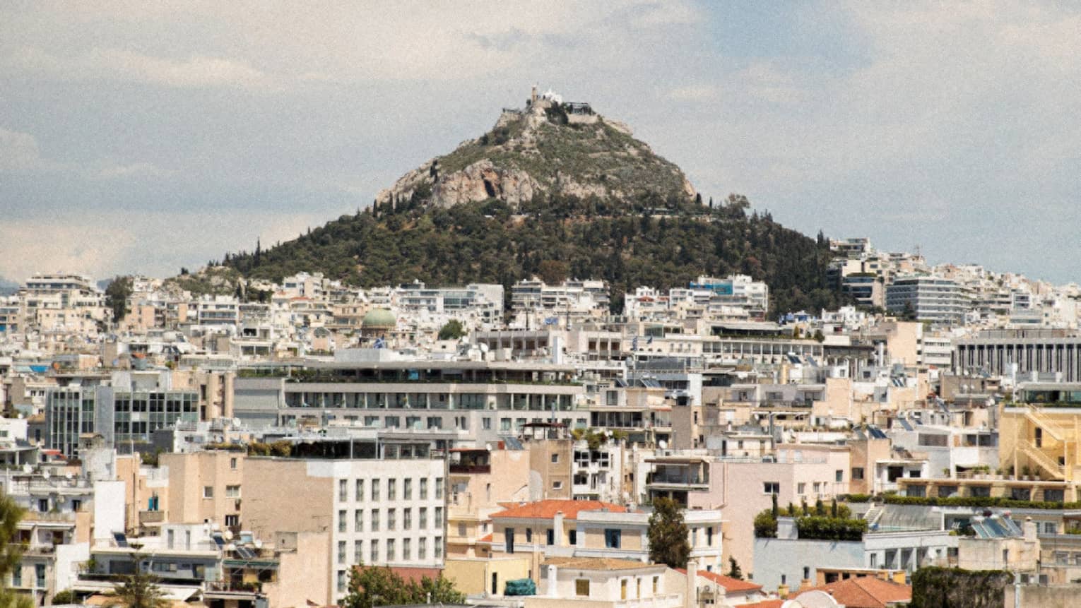 Mount Lycabettus rises from a base of pine trees within the city of Athens, a sprawl of pale modern buildings.