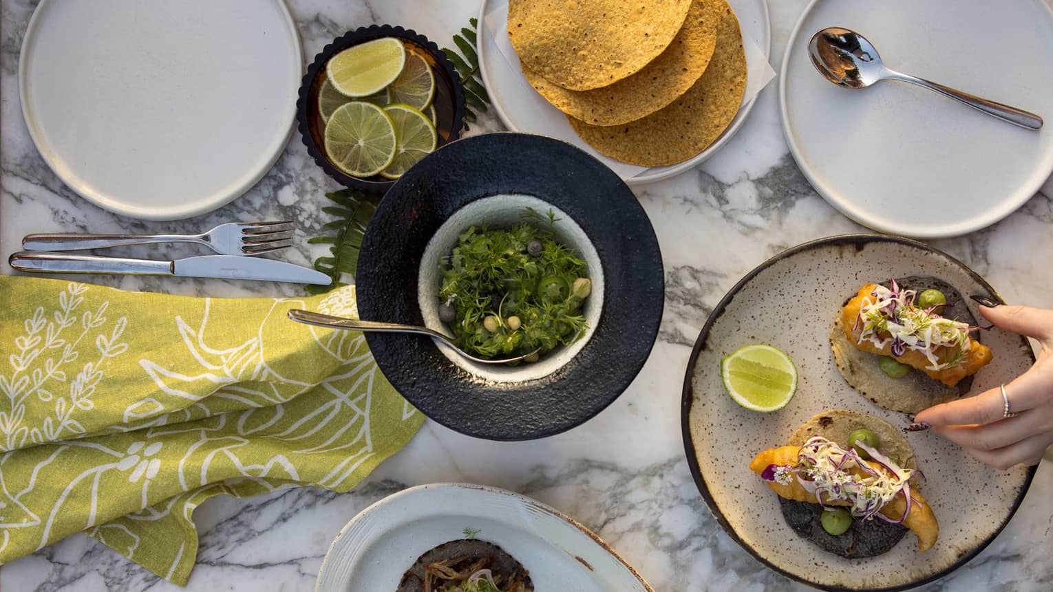 Overhead view of white marble table filled with plates of tacos, bowls of salsa, slices of oranges and limes, with two hands holding cocktails and another set of hands reaching for food