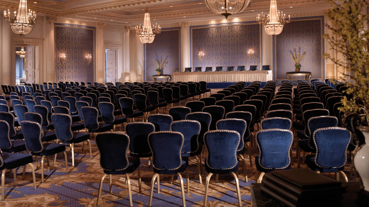 Ballroom with royal blue chairs in rows facing podium with table, textured blue panels, chandeliers 