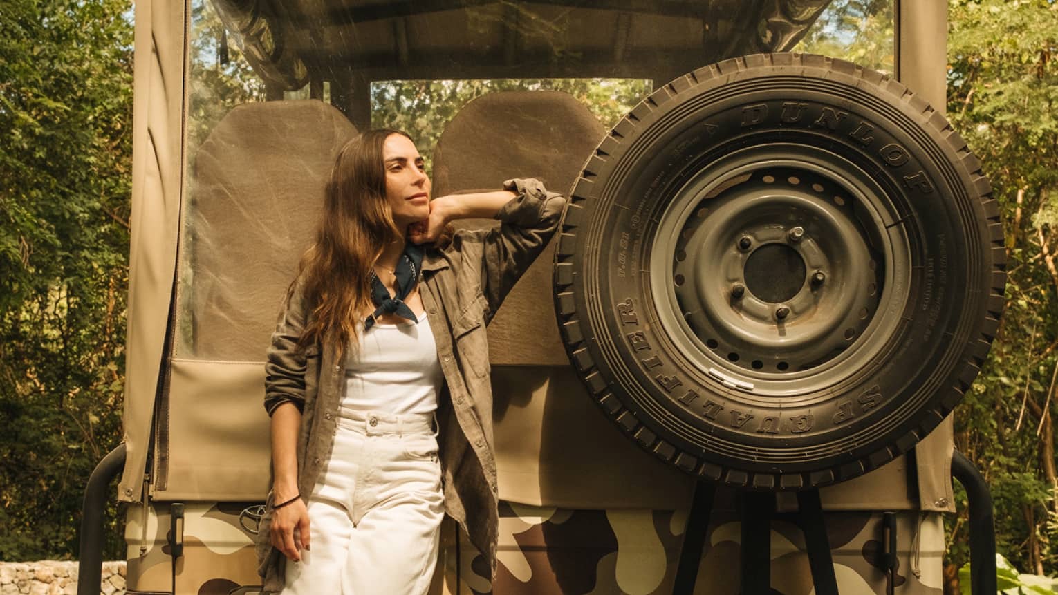 Woman wearing white pants, white shirt and green overshirt stands on the back of an off-road jeep