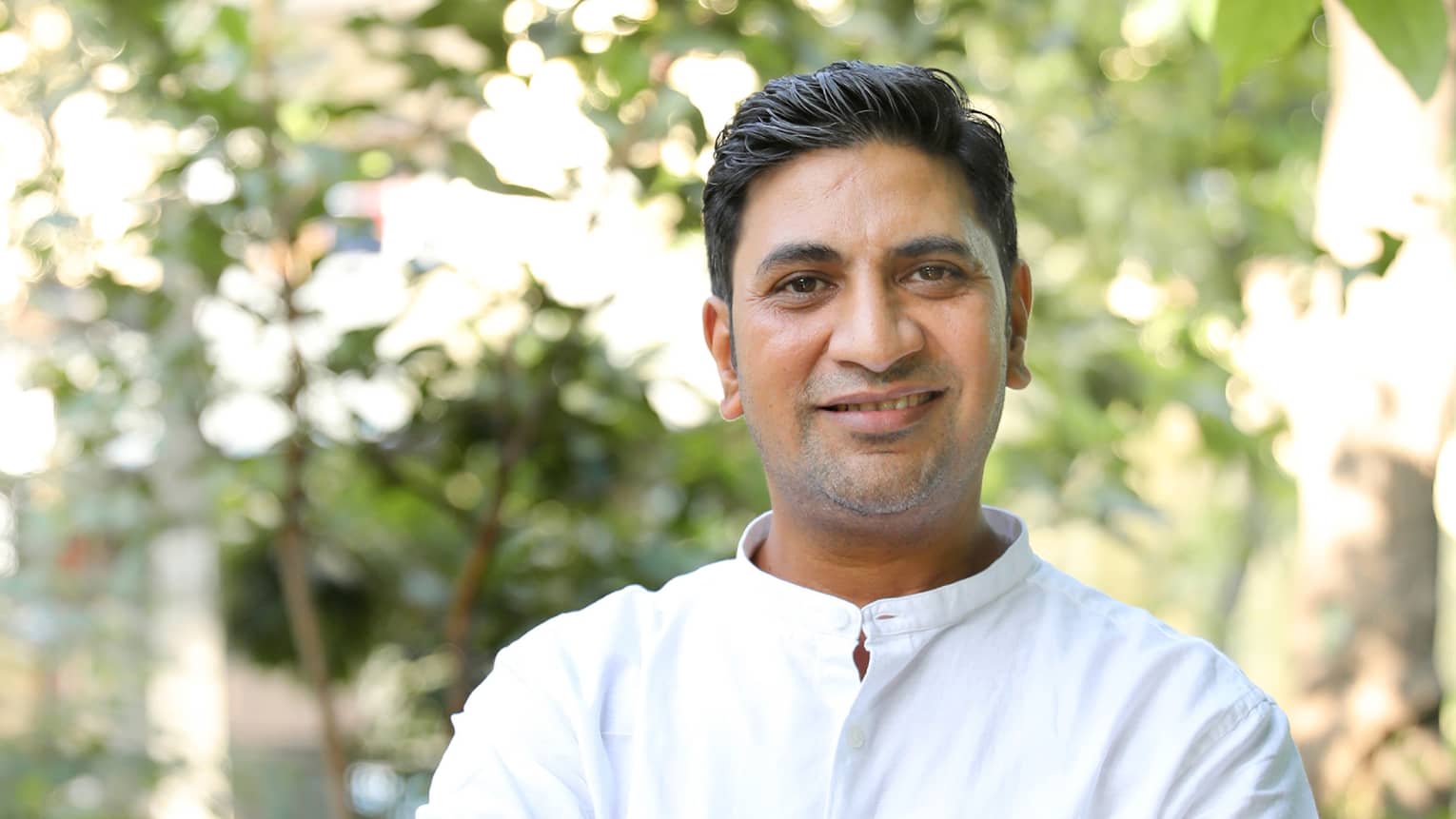Smiling person in long-sleeved white linen shirt stands outdoors with arms crossed