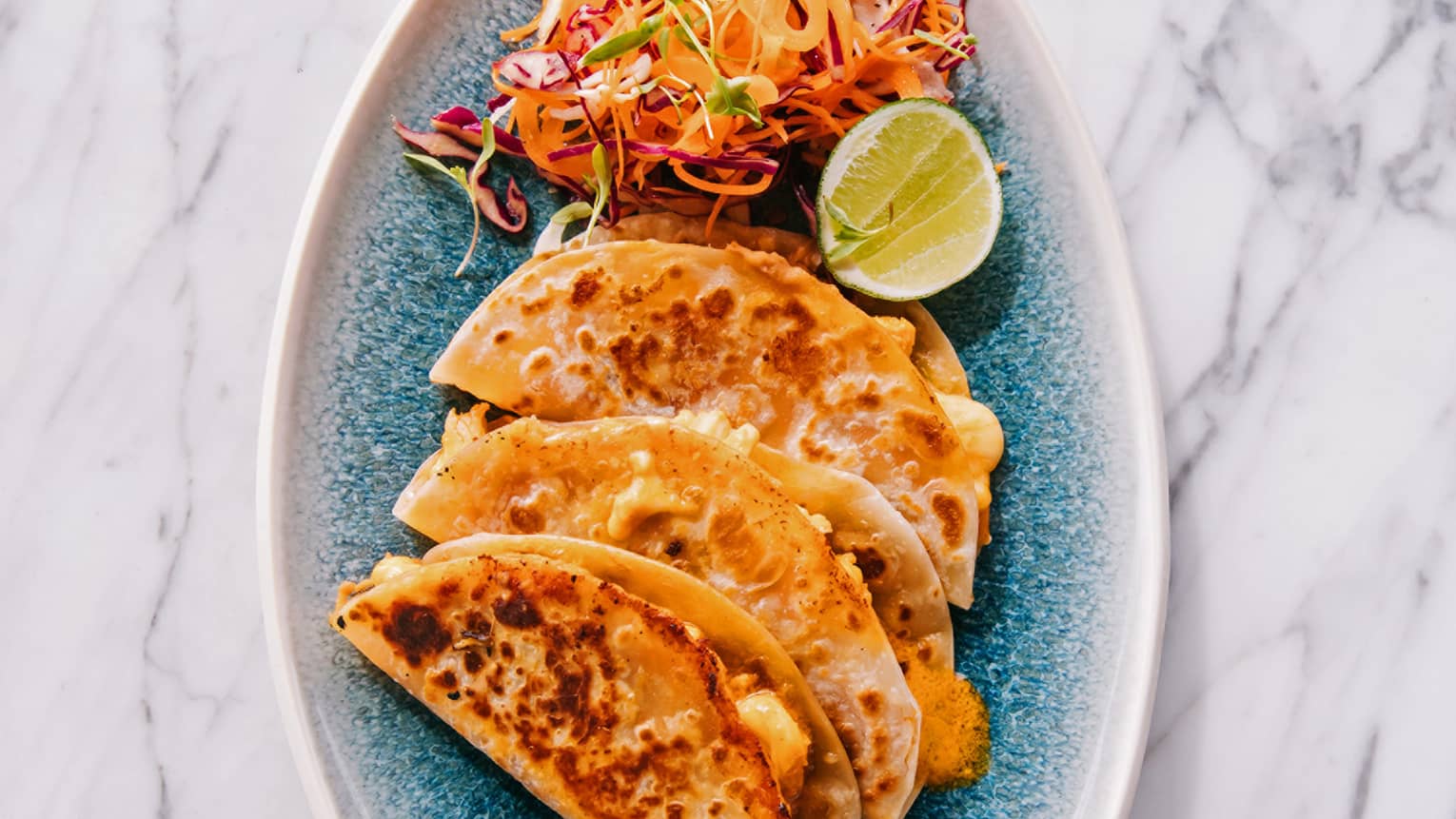 Three tacos and a brightly colored salad with a slice of lime on a blue plate create a visually appealing and appetizing dish.