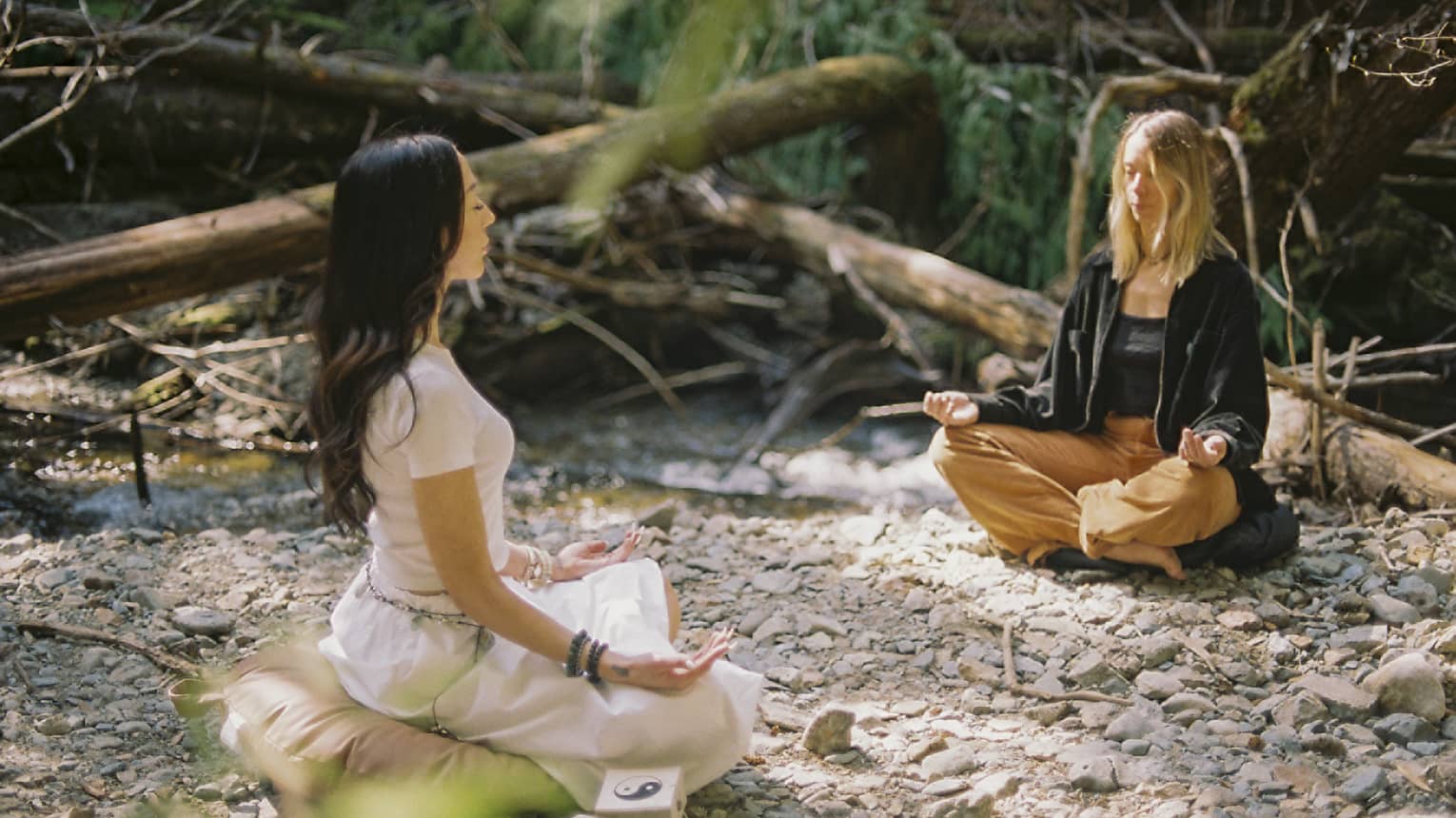An instructor and a guest sit on yoga cushions as they peacefully meditate in a small clearing beside a brook in a forest.