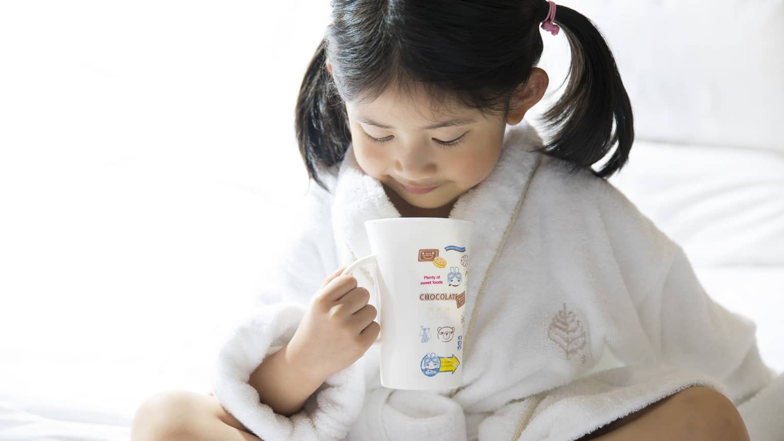 Young child in white bathrobe with hair in ponytails sits cross-legged on bed, holds mug of hot chocolate