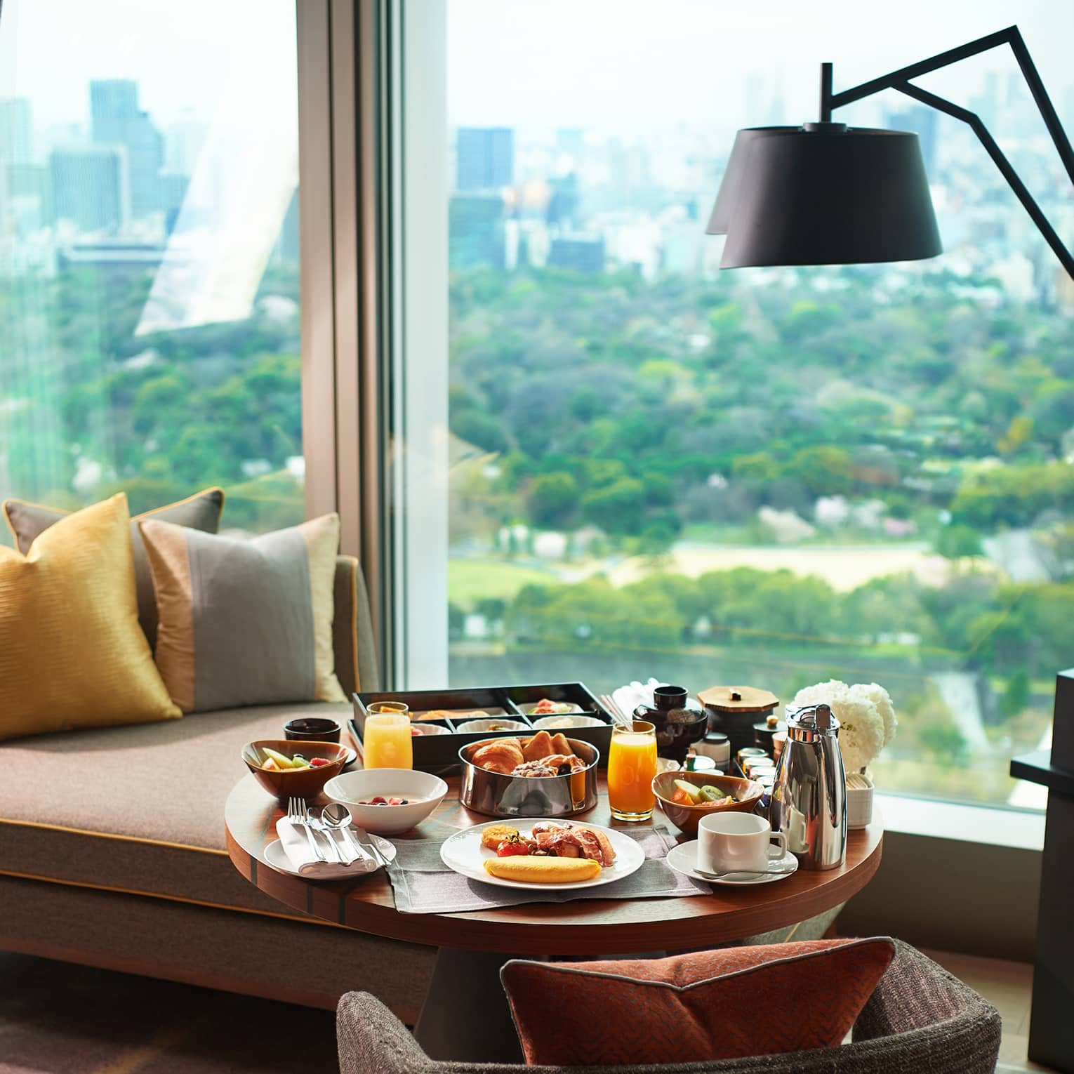 A breakfast tray of pastries, fresh fruit, juice and coffee on a guest room lounge chair at a window overlooking lush gardens.