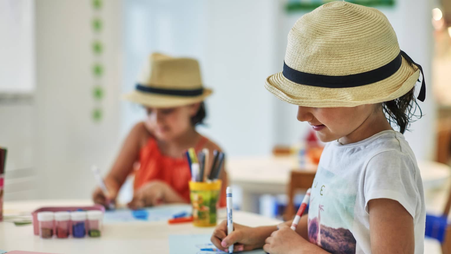 Two smiling children wearing straw fedora hats colour pictures with markers at craft table
