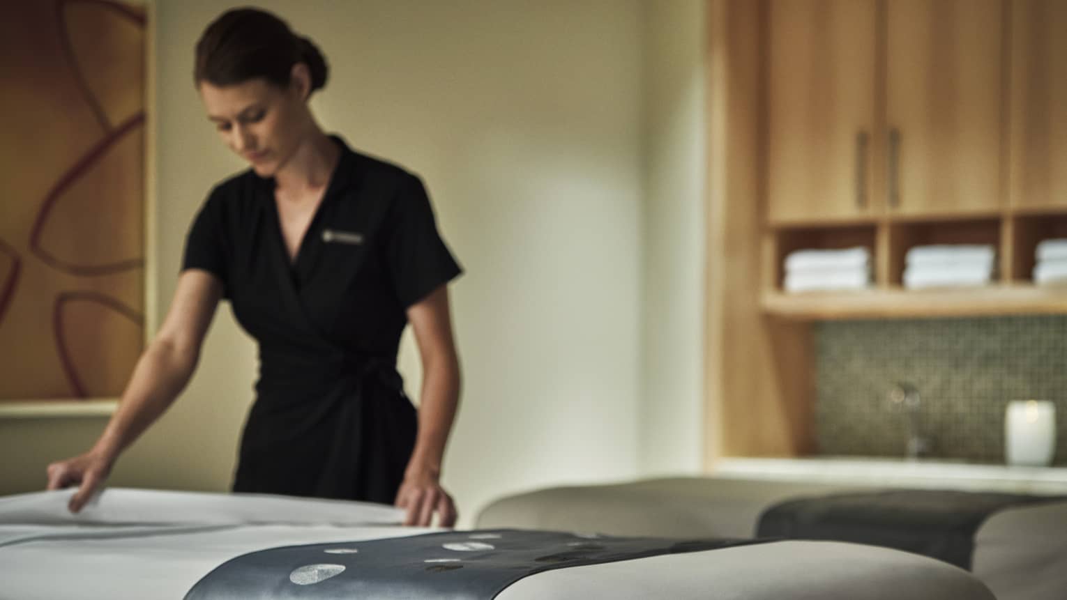 Spa attendant in black dress folds down white sheet on massage table