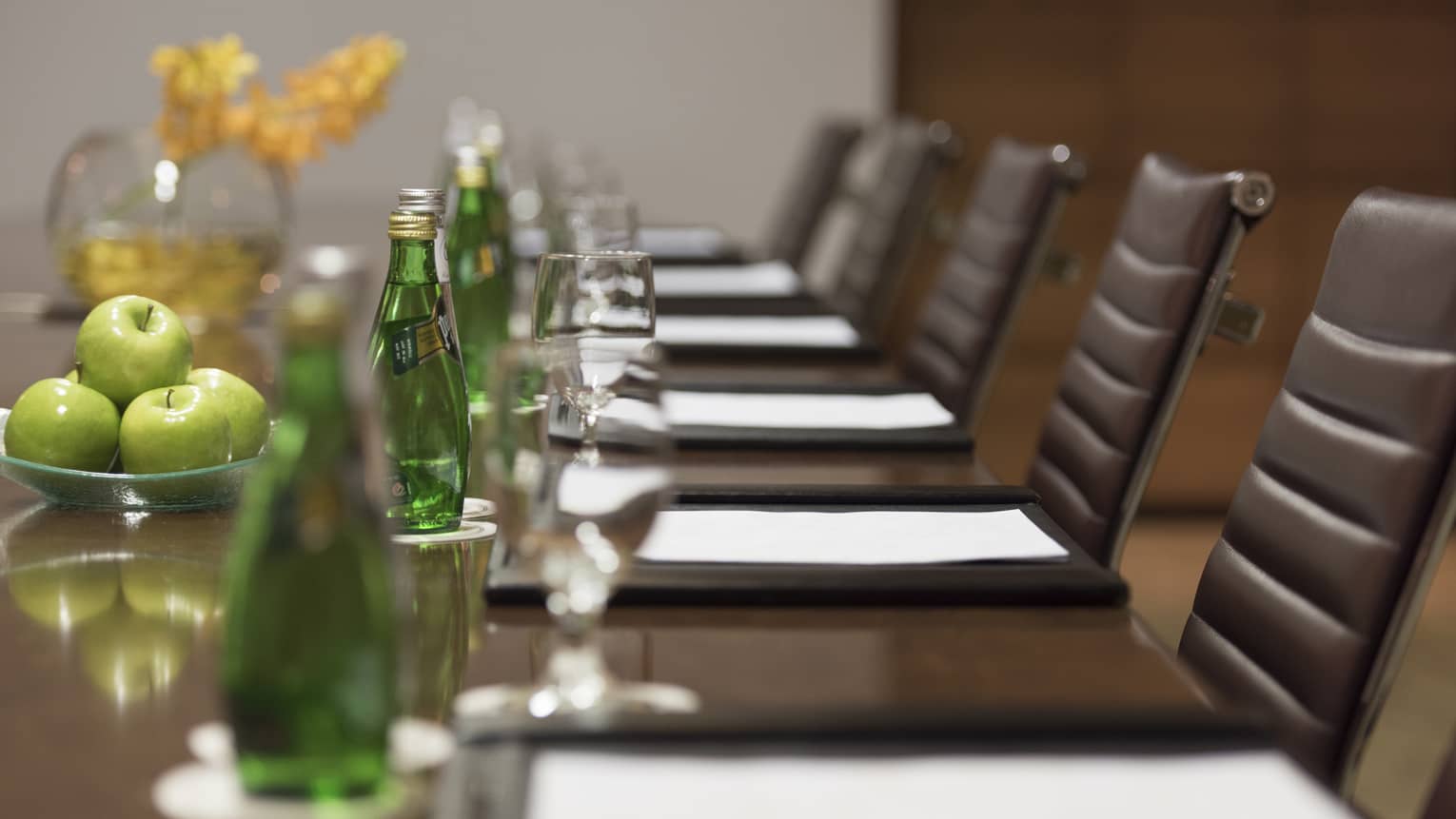 Close-up of business meeting table row of chairs, paper agendas, green glass bottles, apples