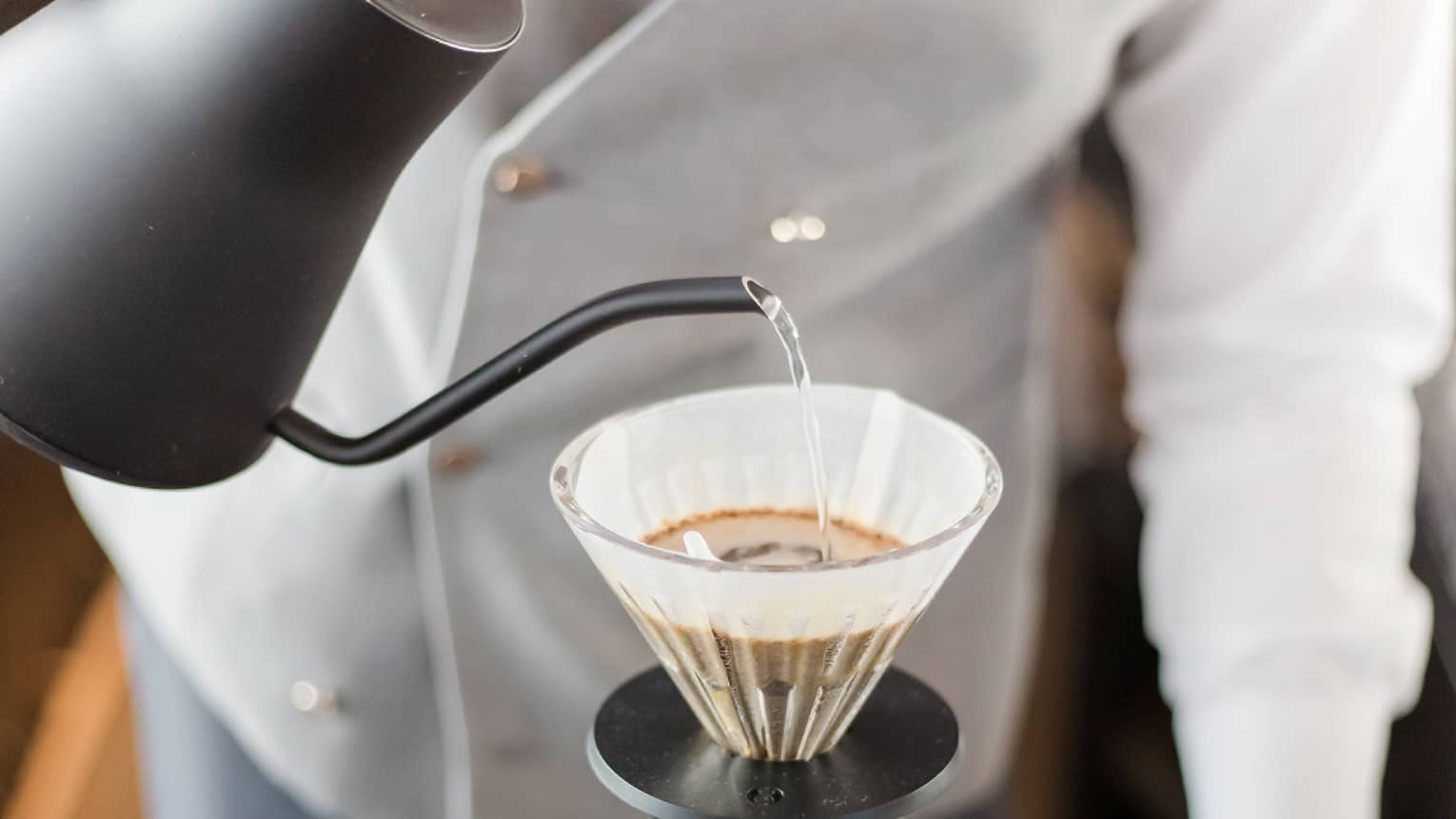 Barista holds black teapot, pours hot water over coffee in cone filter and glass jug on tray