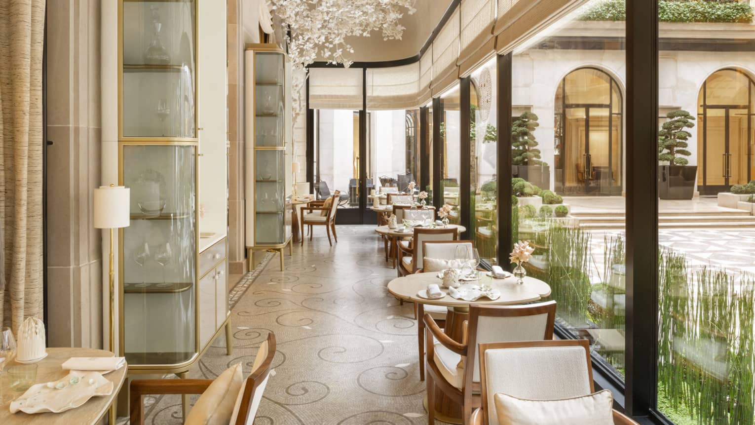 Elegant restaurant corridor-style two-top round tables lining wall of tall windows looking out to courtyard