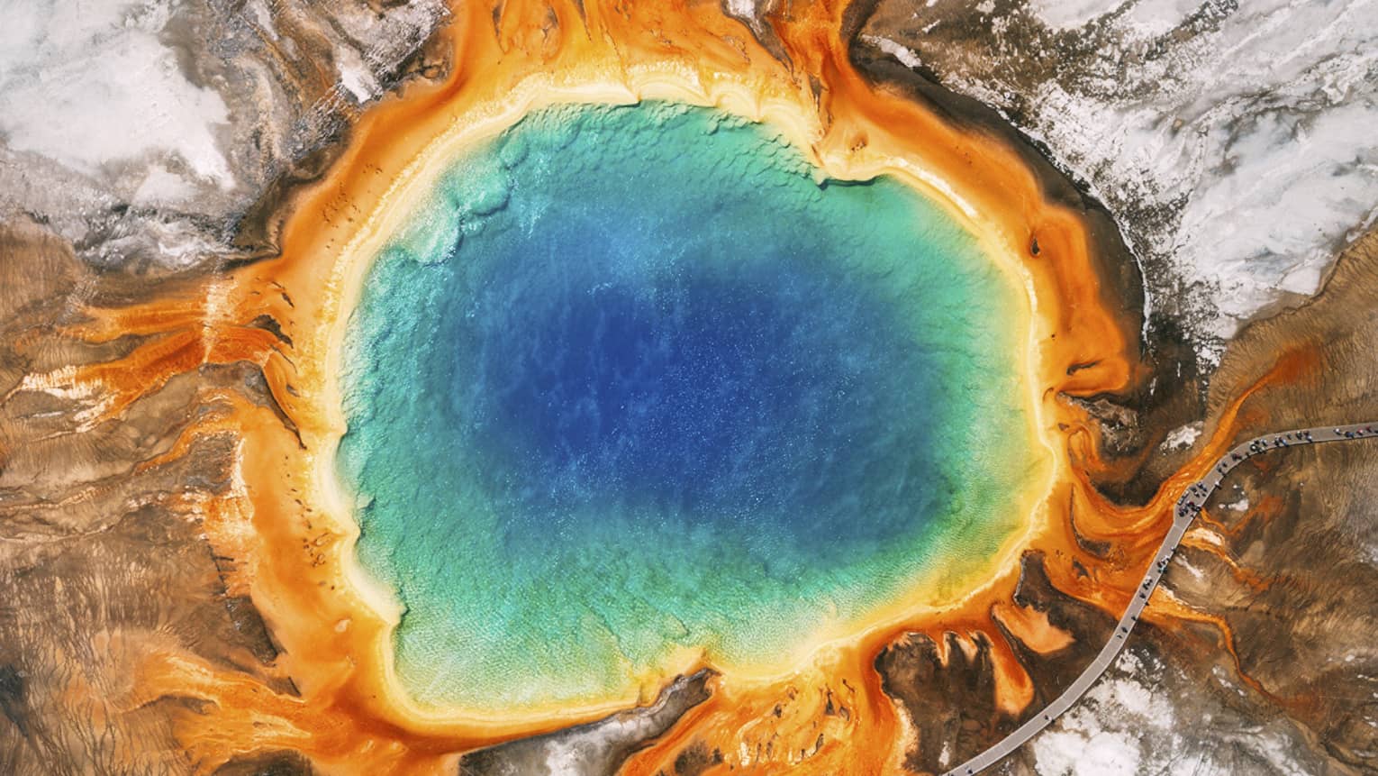 Aerial view of the Grand Prismatic Spring in Yellowstone, featuring vibrant blue, green, yellow and orange concentric rings, surrounded by rocky terrain