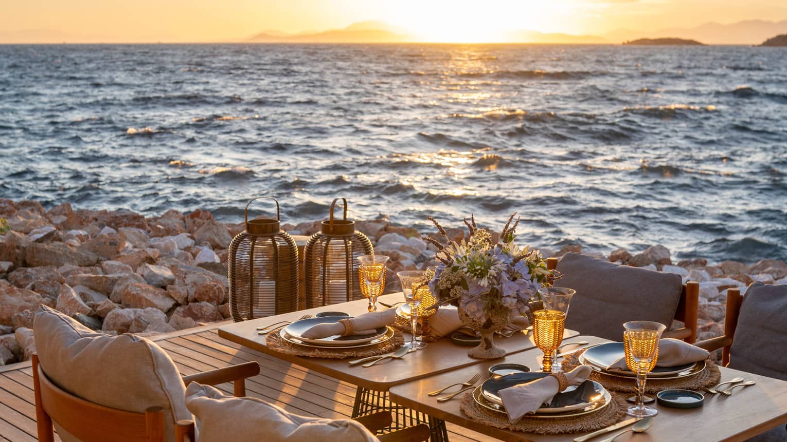 A table laid with sparkling glasses and a bouquet of flowers set on a deck on the rocky shore, the sunlit waves metres away.