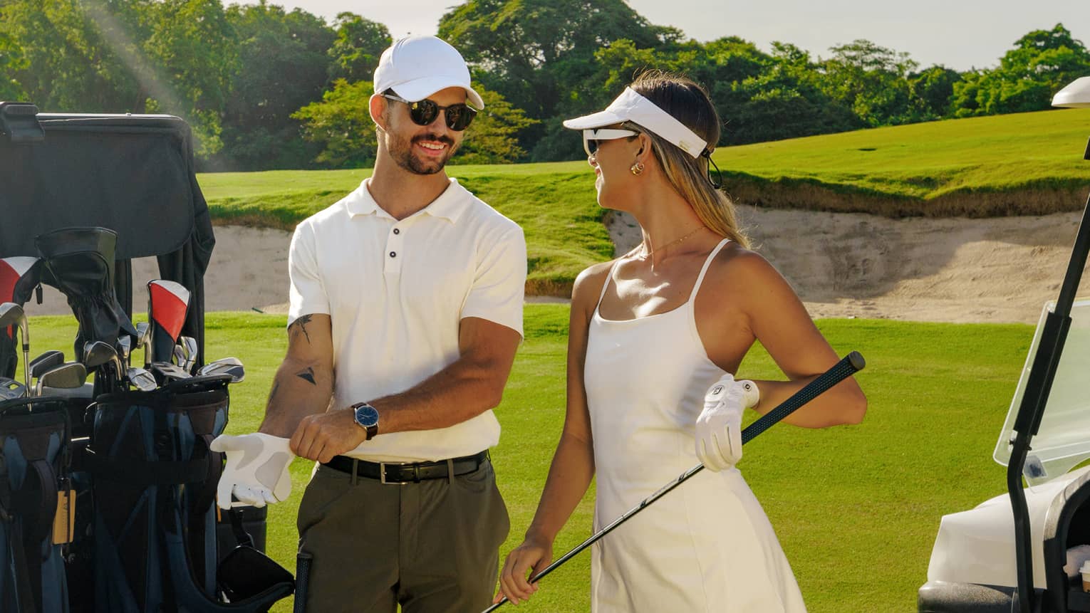A couple wearing white golf attire stand together next to a golf cart in the midday sun