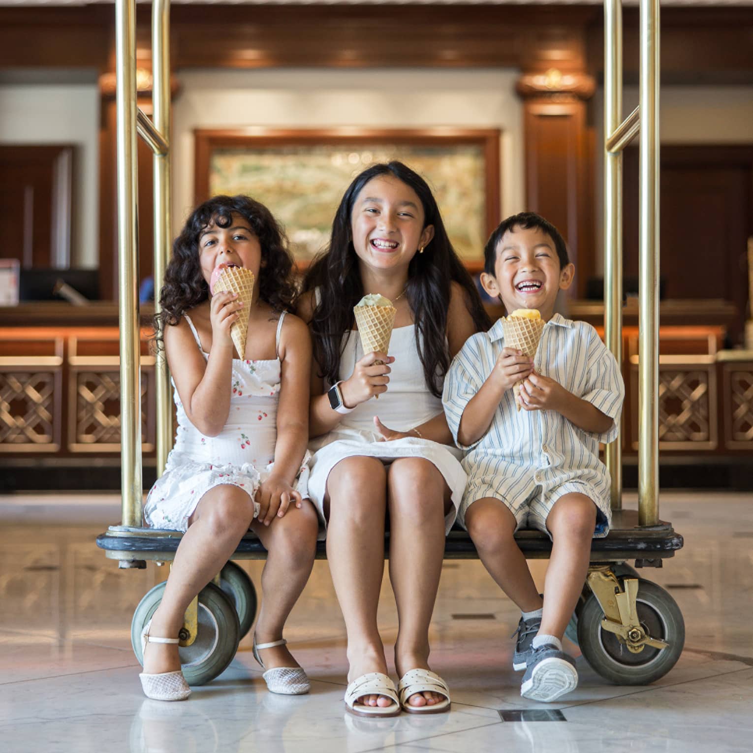 Three children sit on a gold hotel luggage cart eating ice cream