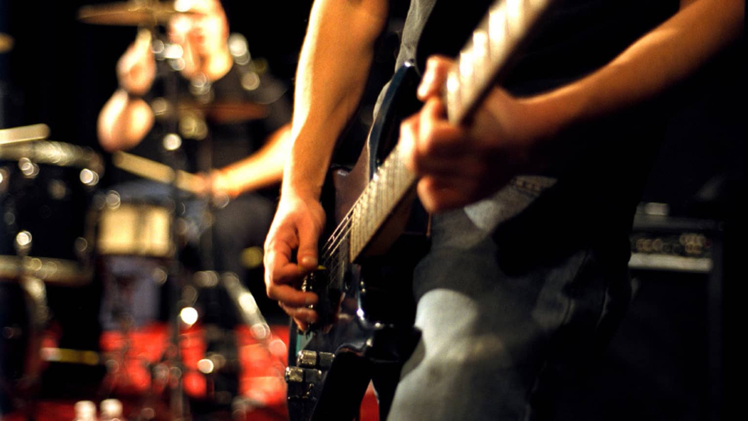 Close-up of musician's arms and hands playing an electric guitar with drumset in the background.
