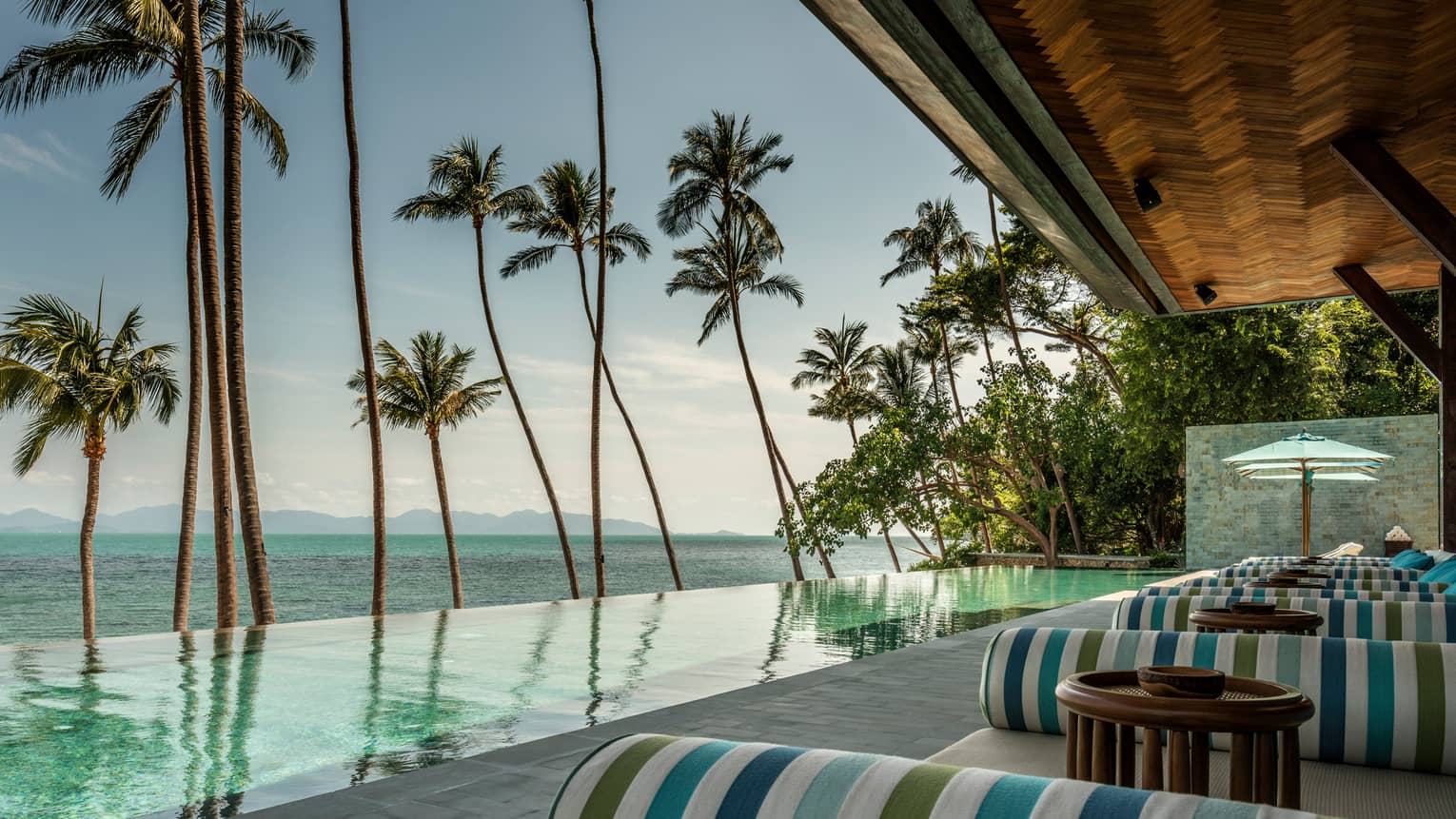 Green-and-blue striped banquettes on patio by infinity swimming pool, ocean