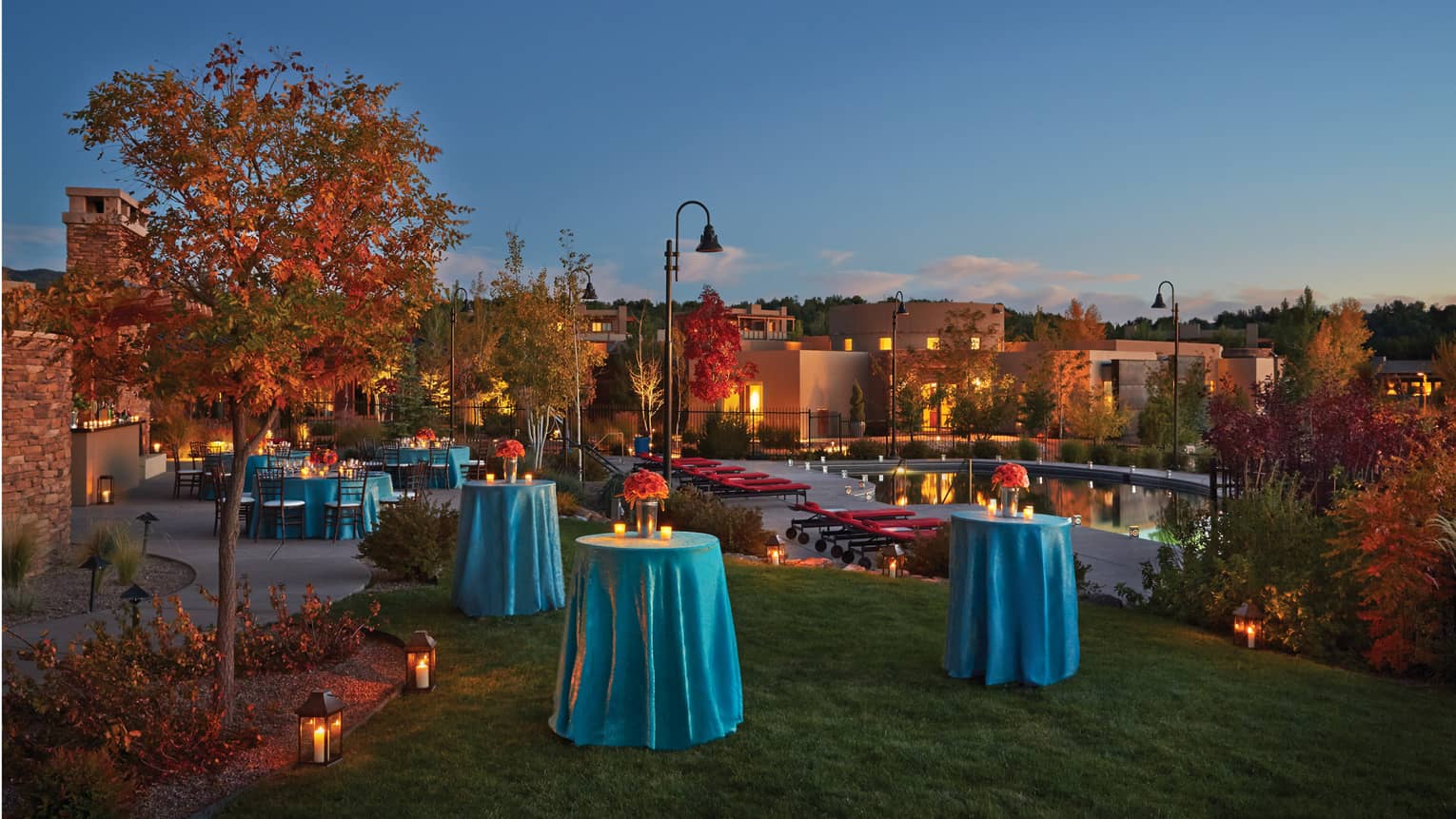 Cocktail tables with blue table clothes and flowers on Rows of chairs on lawn by pool at dusk 