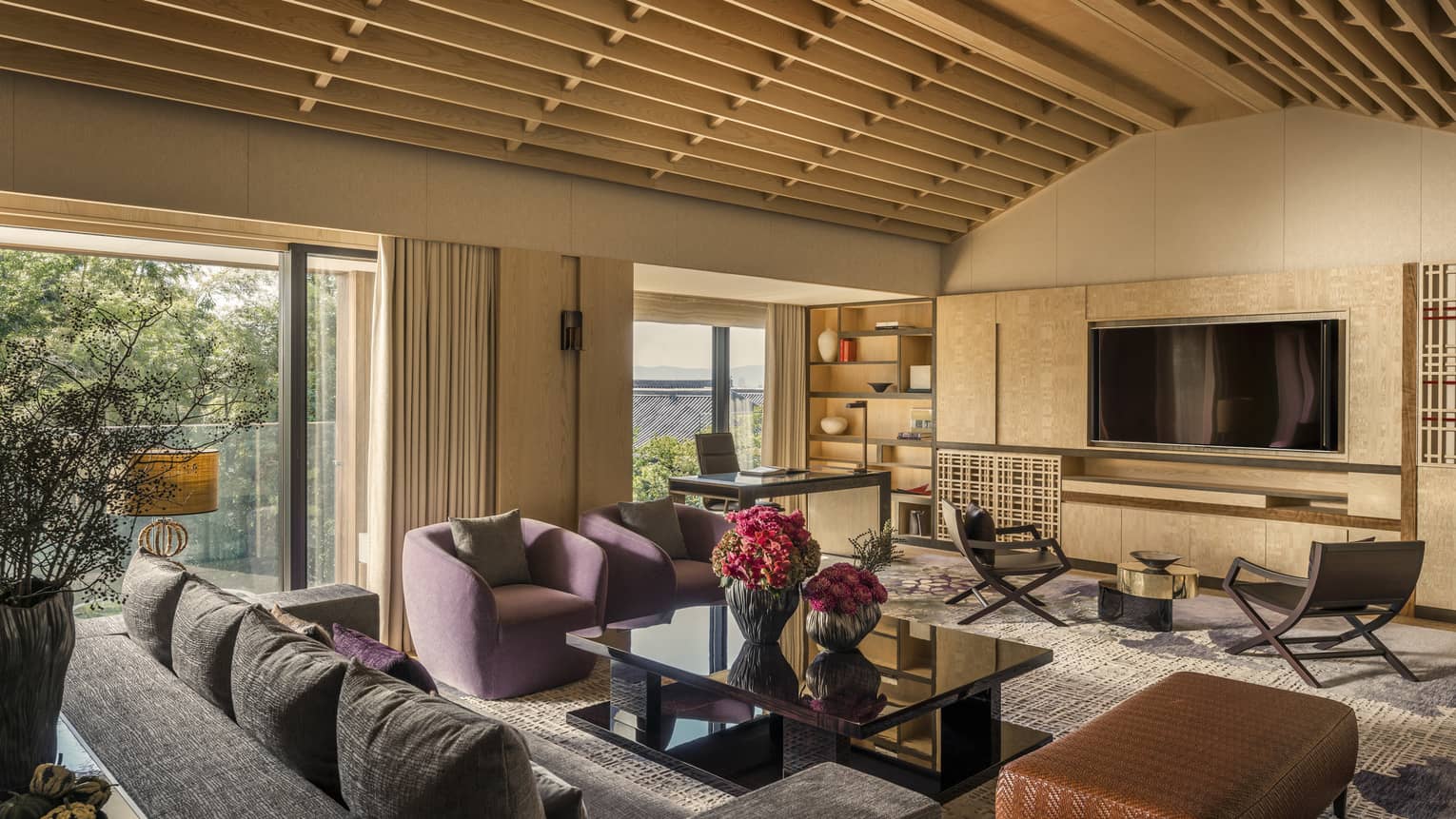 Living area with vaulted ceiling, purple armchairs, sofa, black coffee table, wall-mounted TV, desk, chairs and large windows overlooking greenery