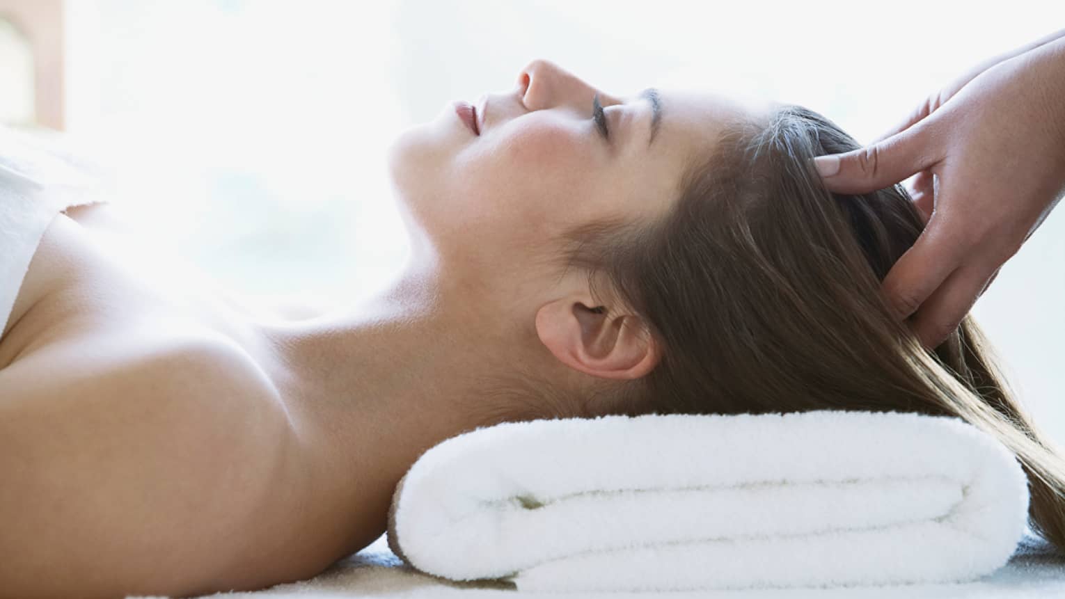Woman lays on back with head on folded white towel, closes eyes as hands massage her scalp