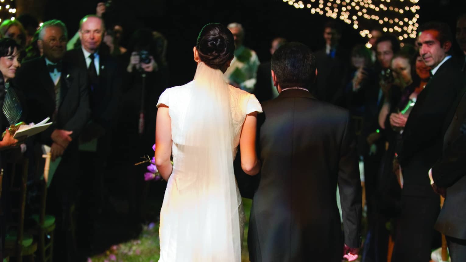 Back if bride wearing long gown walking down aisle at night, tree with lights