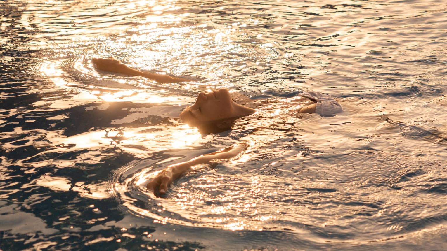 A person floating on their back in the ocean, with sunlight rippling across the water.