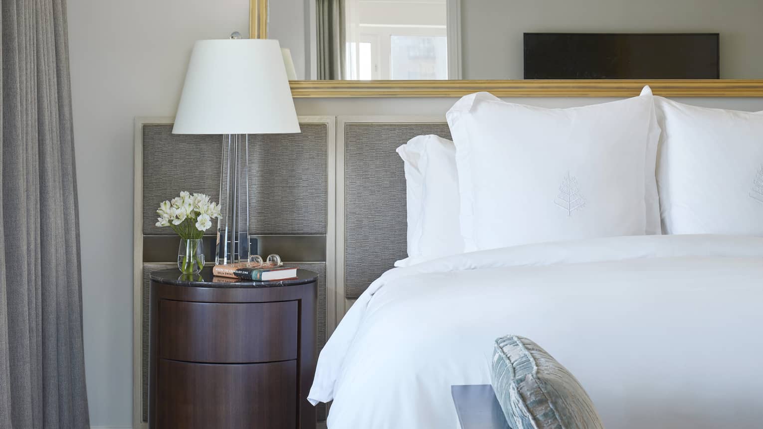 White bed with grey headboard, brown nightstand with lamp, flowers, book and eyeglasses