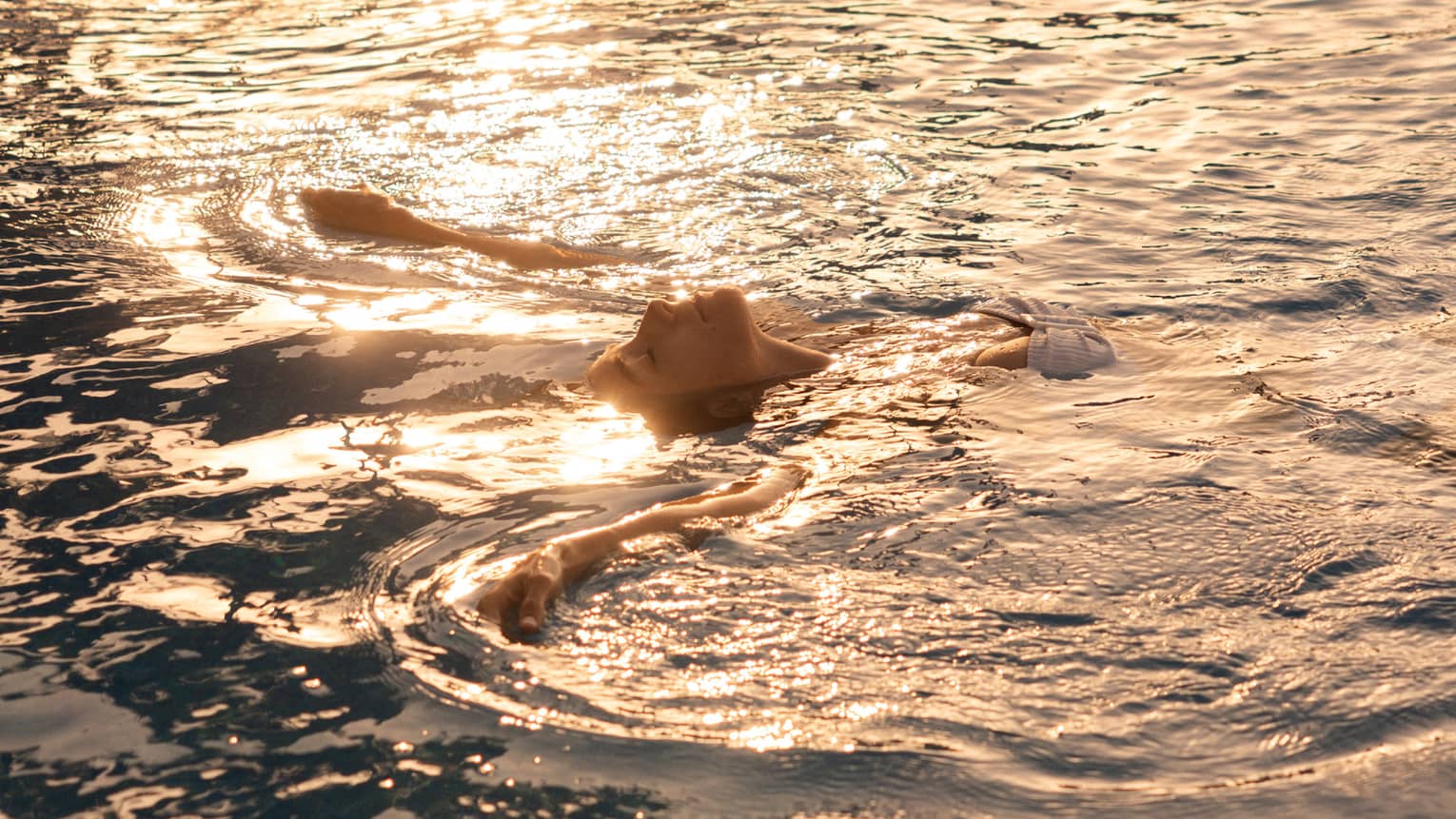 A person floating on their back in the ocean, with sunlight rippling across the water.